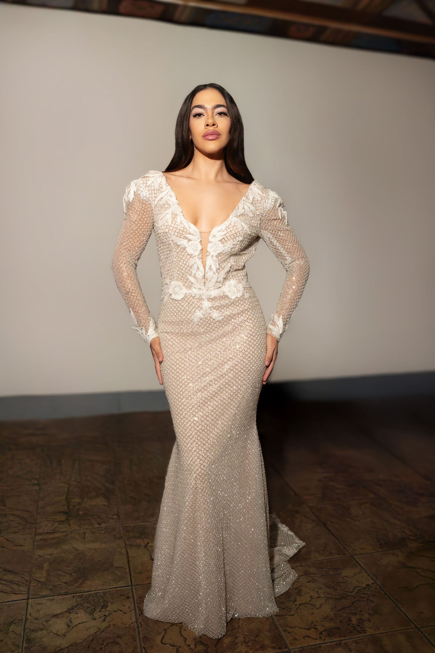 A model poses in a sheer beige lace dress with long sleeves against a studio backdrop.
