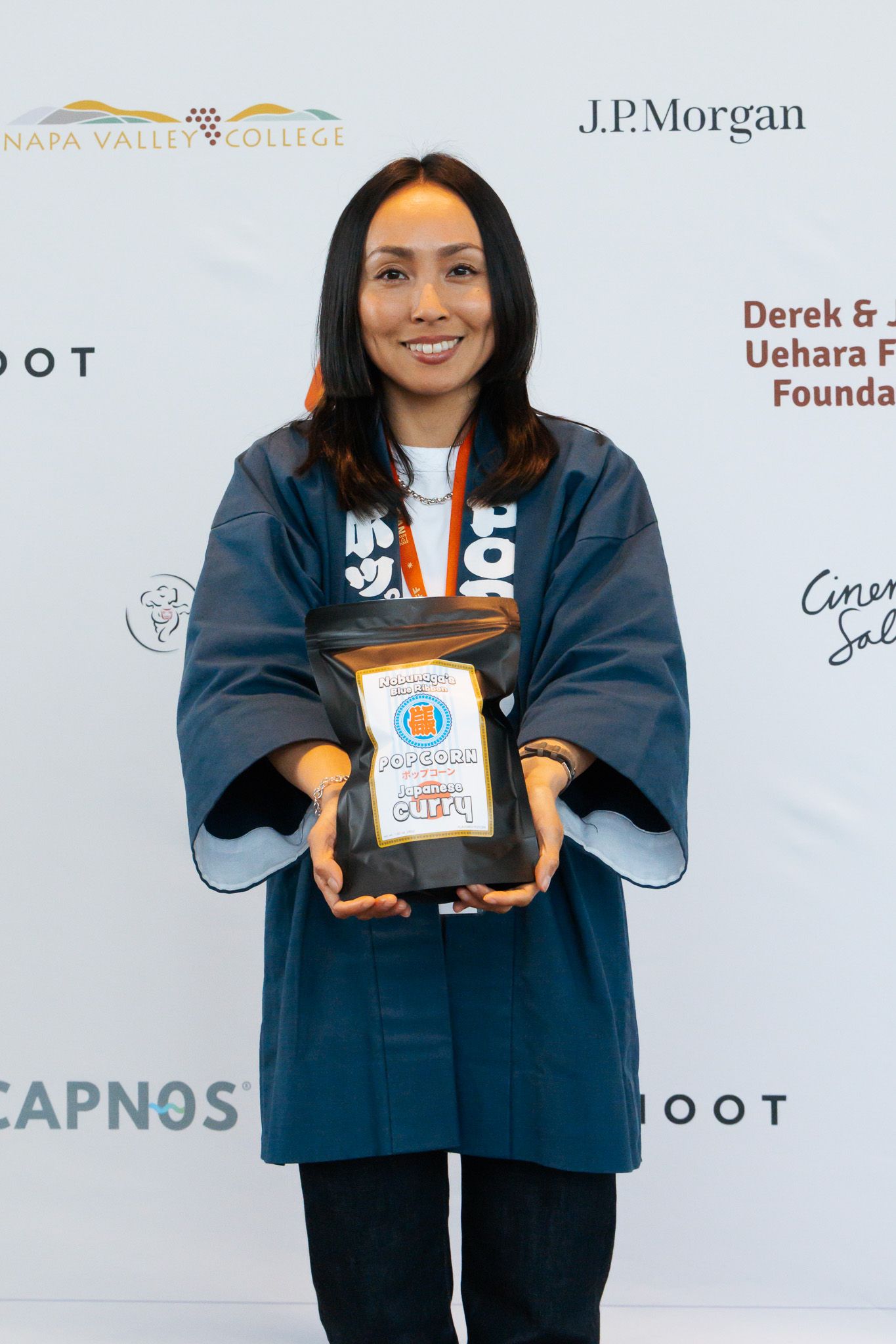 Three award recipients in matching navy jackets hold up trophies at a ceremony with sponsor logos displayed on the backdrop.