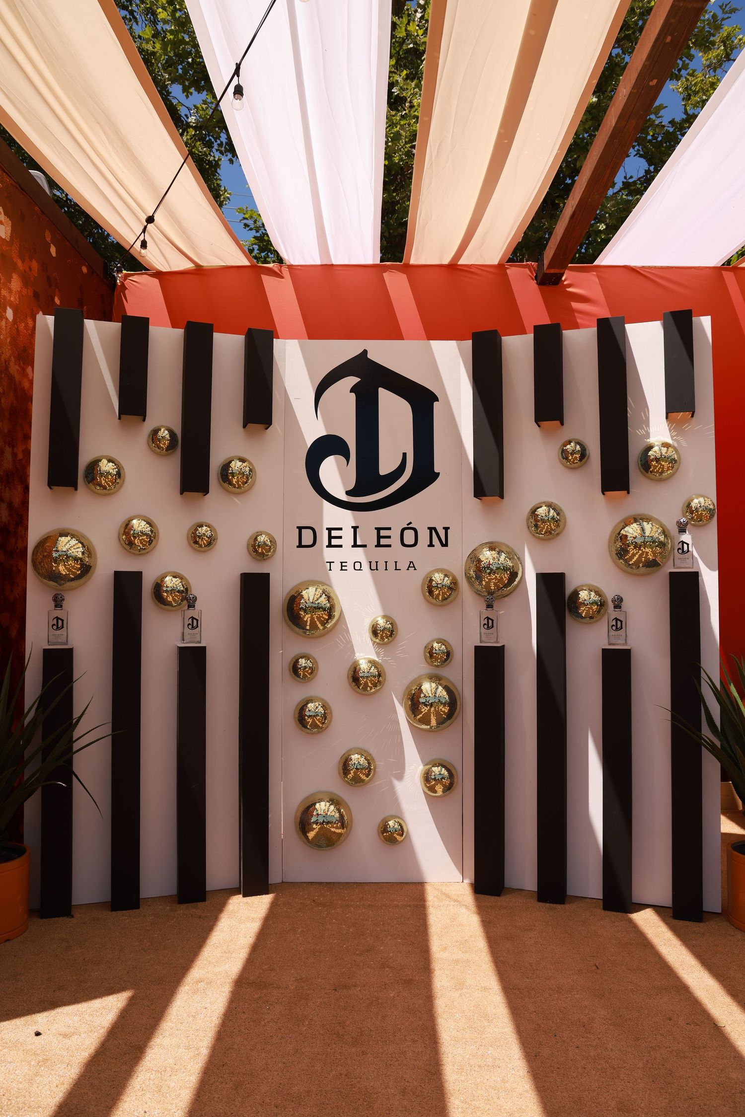 Decorative gold orbs adorn a black and white striped wall under a white pergola at a DeLeon tequila event.