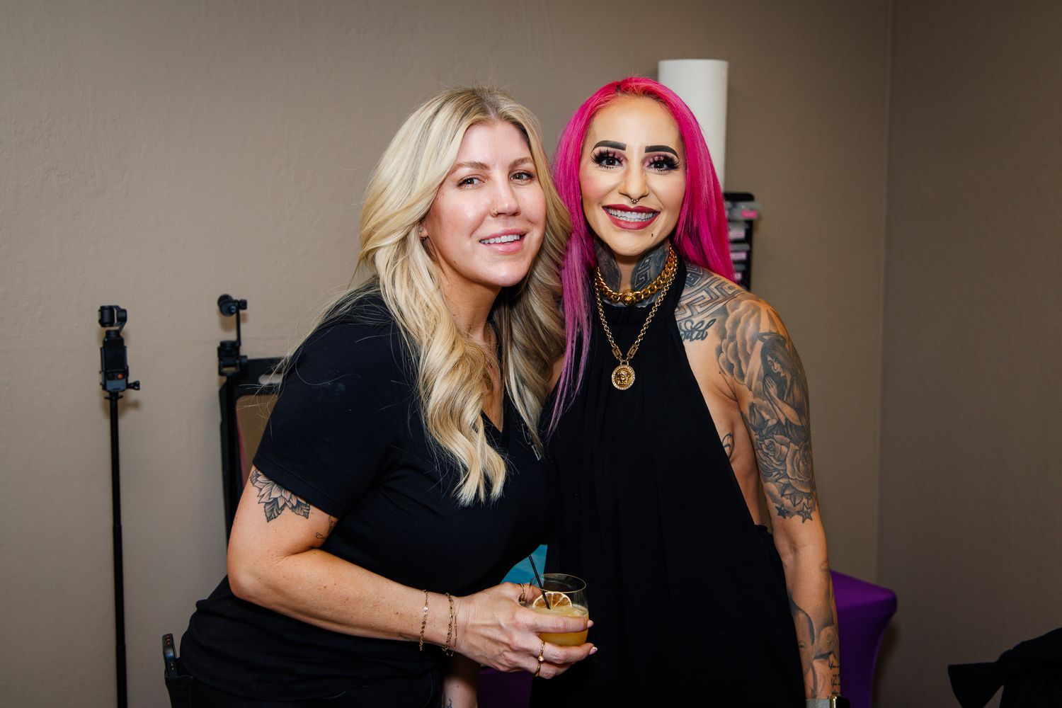 Two women smiling at event, one with blonde hair and one with pink hair and tattoos, holding drinks.