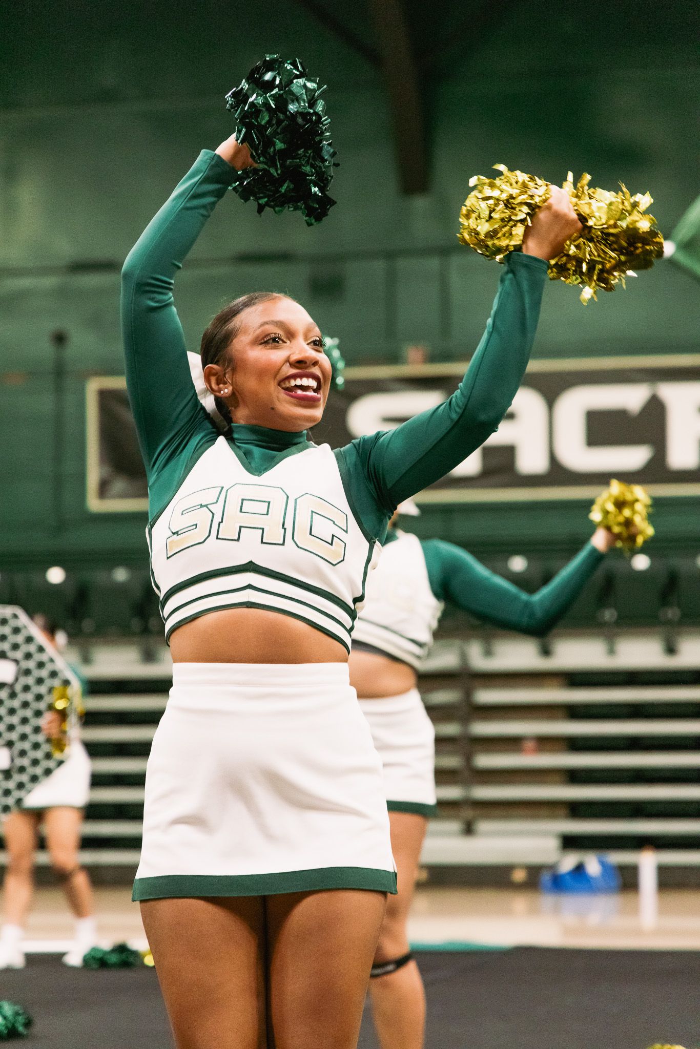 SAC cheerleader in green and white uniform raises gold and green pom-poms while smiling during a performance.