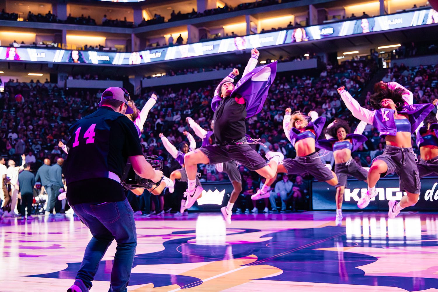 NBA arena dance team performs energetic halftime show on court with purple Kings logo, crowd watching under bright lights.