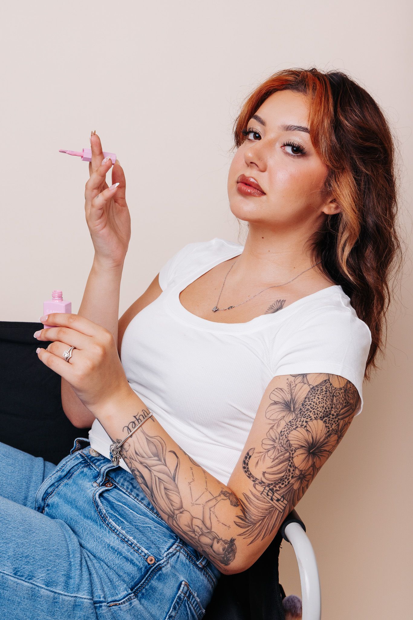 Young woman with tattoos on her arm holding pink nail polish, wearing white crop top and jeans.