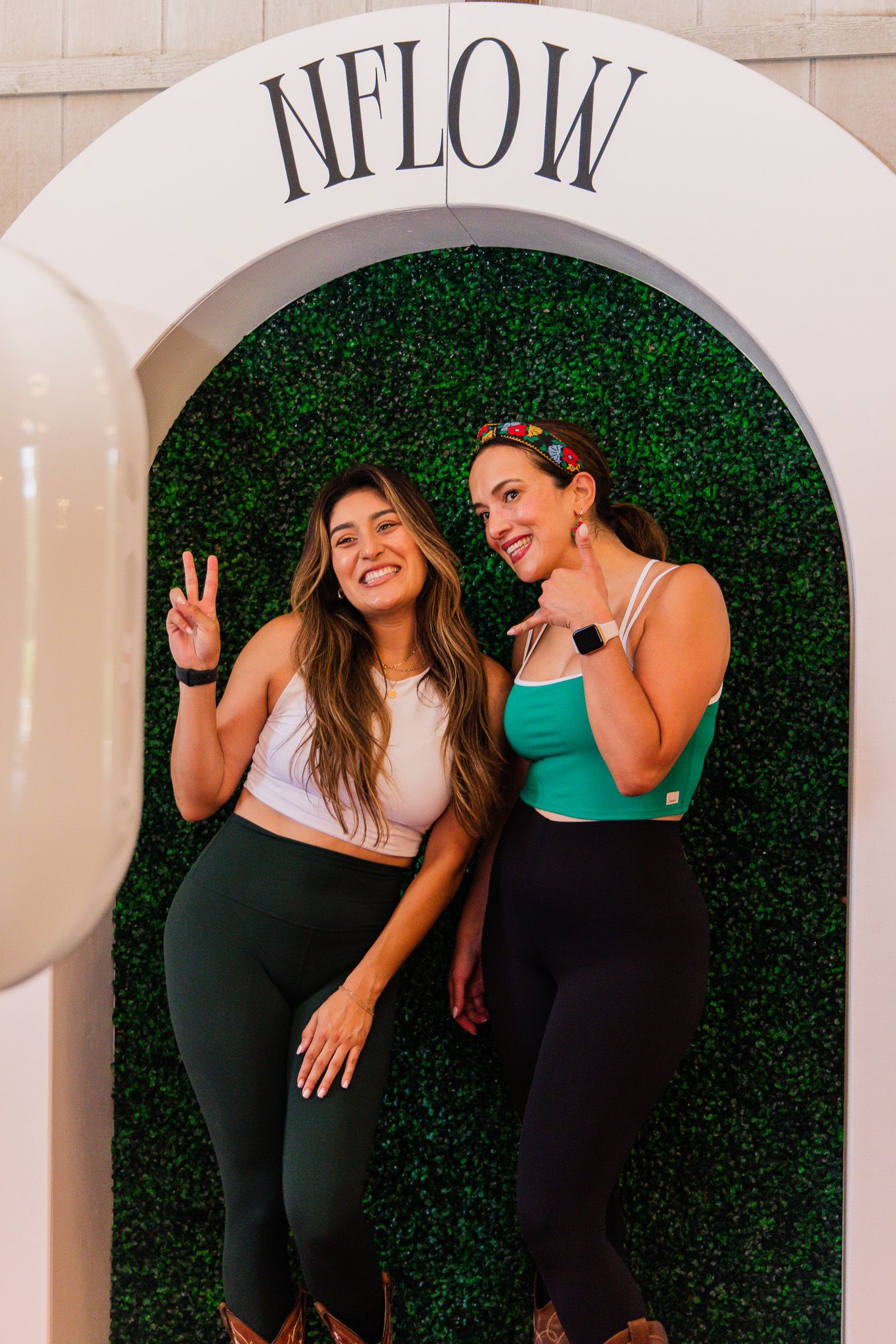 Two women pose and smile in front of an NFLOW branded green boxwood hedge arch backdrop.