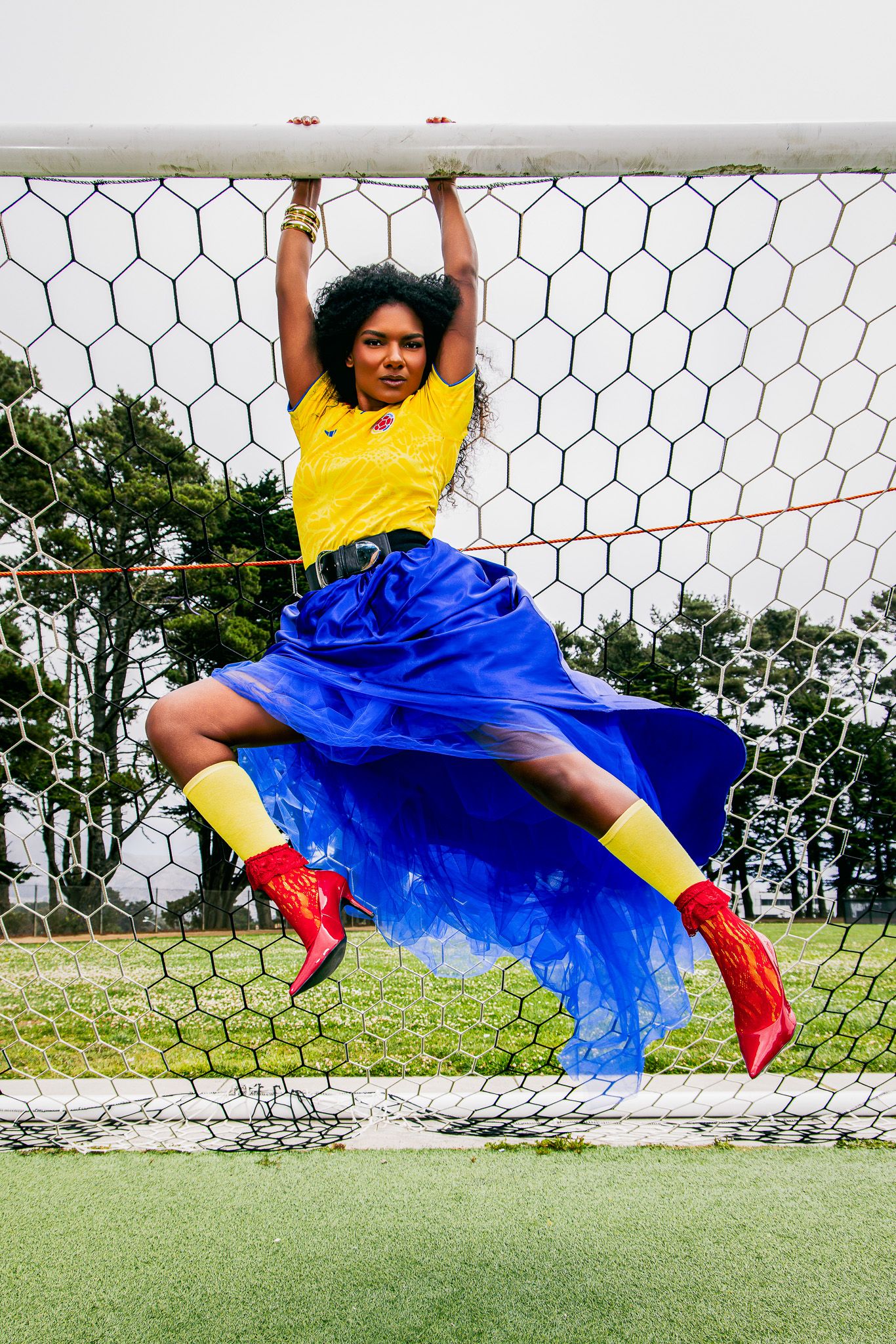 Woman in yellow Colombia jersey, blue tulle skirt, and red boots hangs from a soccer goal post on a green field.
