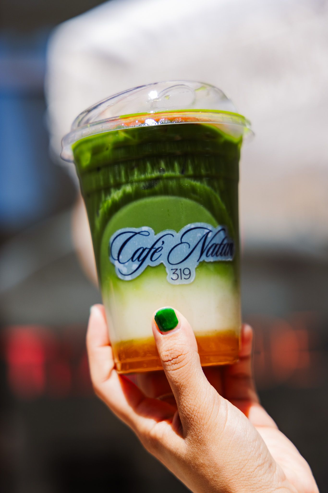 Hand holding Cafe Natur 319 iced matcha latte in clear plastic cup.