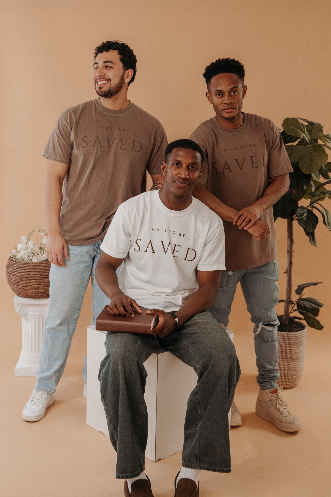 People wearing Saved branded t-shirts in brown and white sit and stand against a beige backdrop with a fiddle leaf plant.