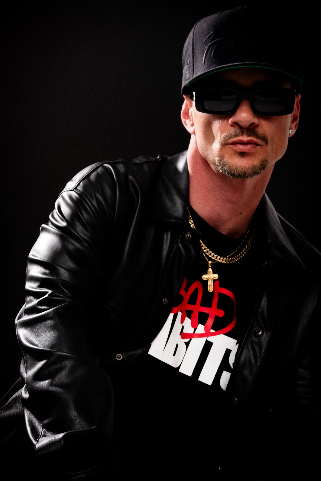 Four dramatic black and white photographs show a figure in dark clothing and sunglasses with a gold chain against a black background.