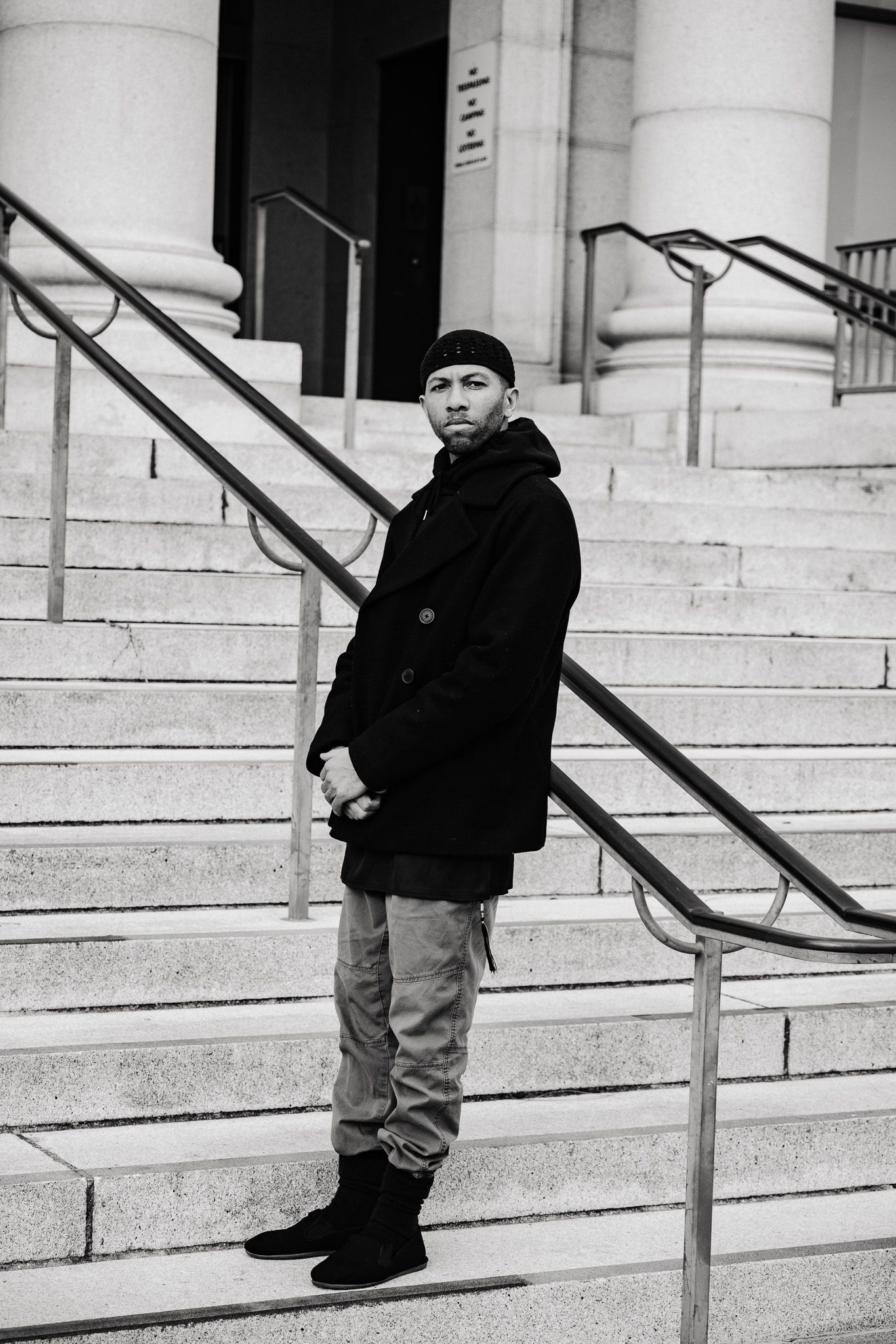 A person in winter clothing stands on concrete steps with metal handrails in a black and white photograph.