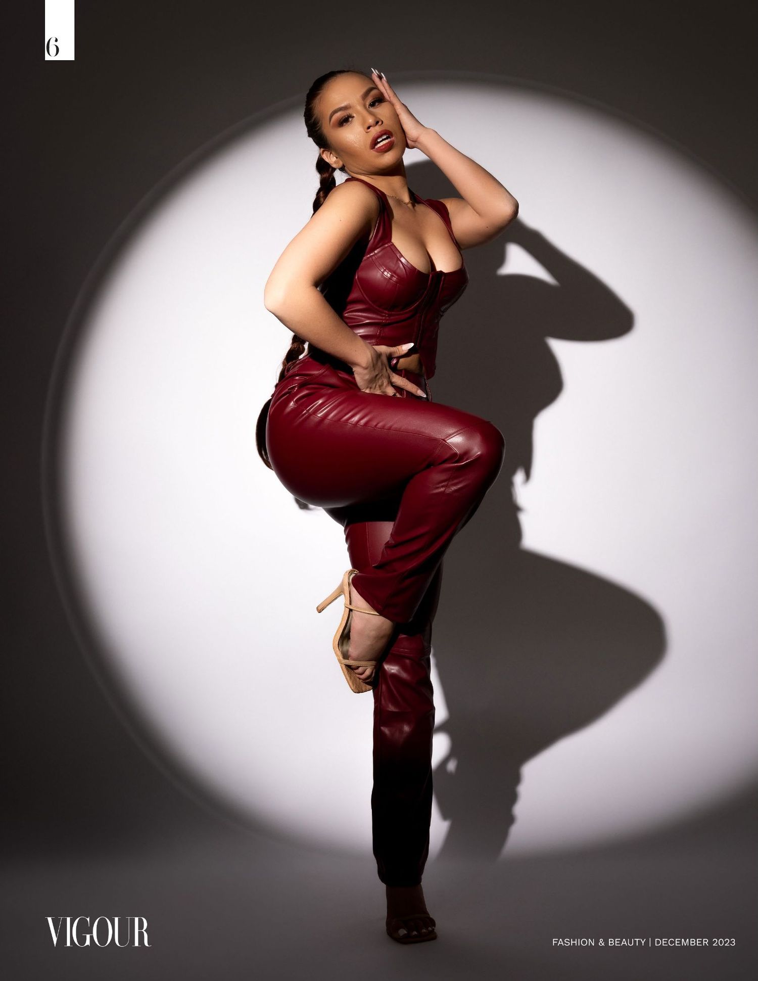 Model poses dramatically in burgundy leather jumpsuit against spotlight creating bold shadow on gray backdrop.