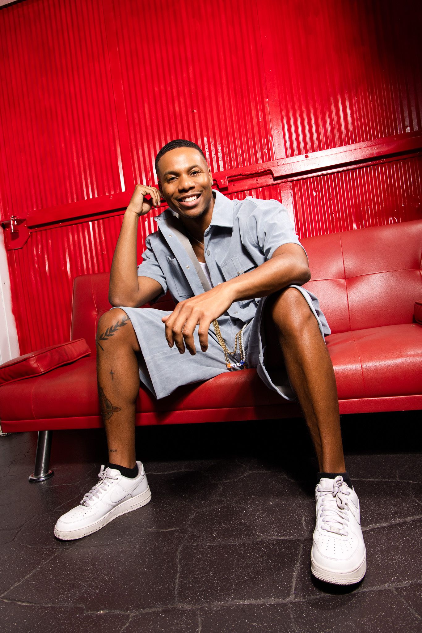 Smiling young Black man in gray shorts sitting on a red leather couch wearing white sneakers against a red wall.