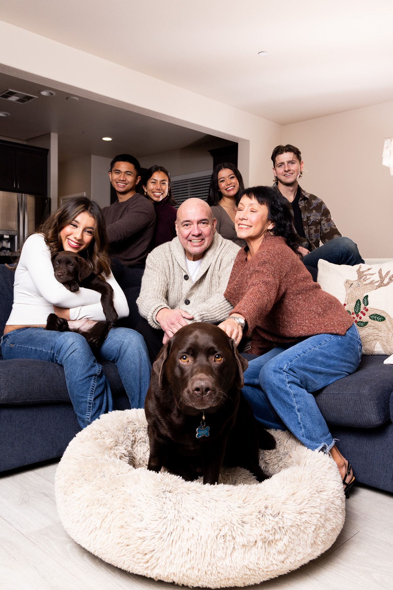 A chocolate Labrador retriever lounges comfortably in a plush round dog bed while family members relax on the couch nearby.