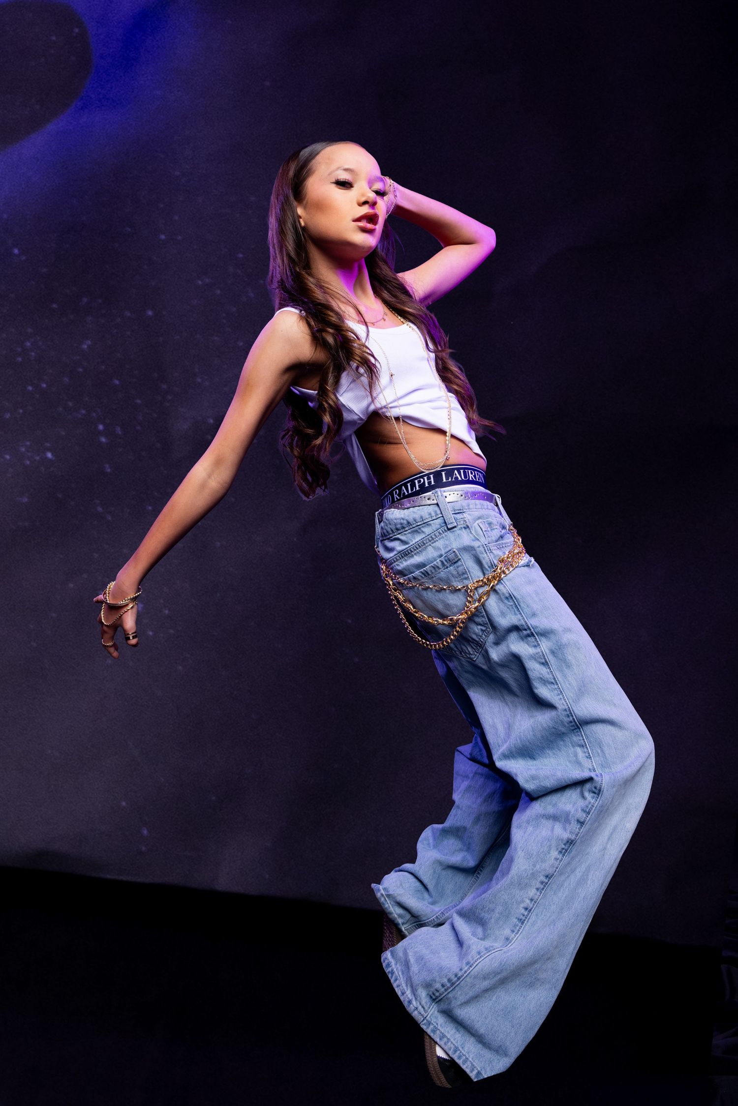 Young woman performing on stage in white crop top and wide-leg jeans with chain belt under purple stage lighting.