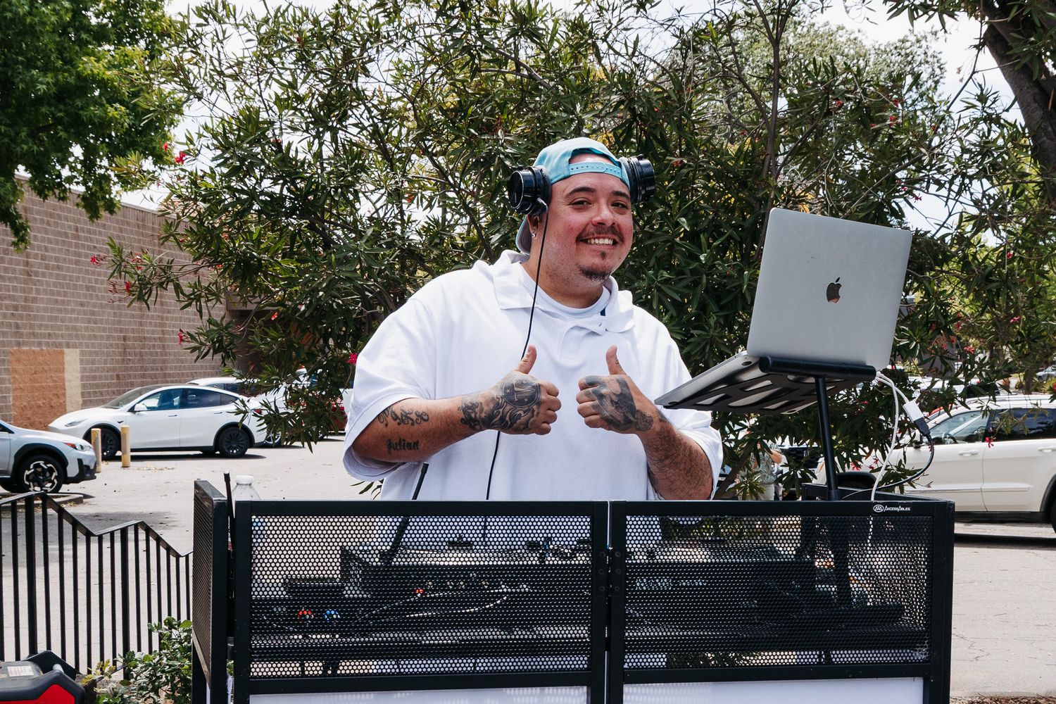 Tattooed DJ smiles holding MacBook laptop outdoors at sunny event with DJ equipment setup.