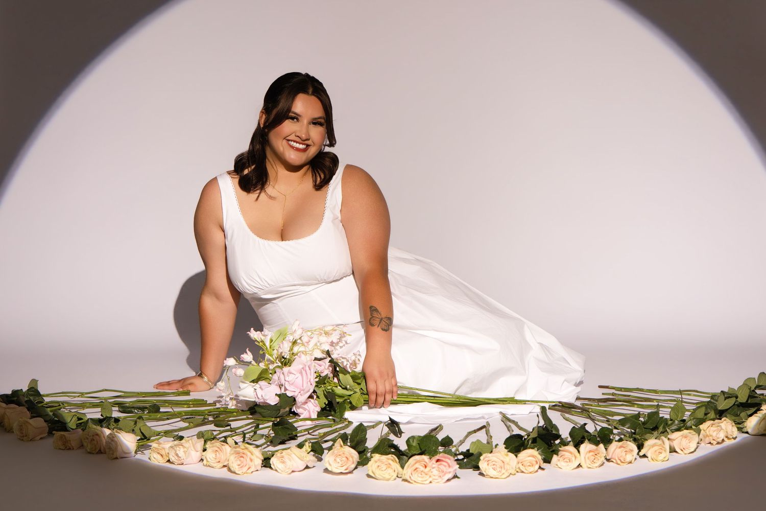 Plus-size bride in white gown seated gracefully among white roses arranged in a circle on a light gray background.