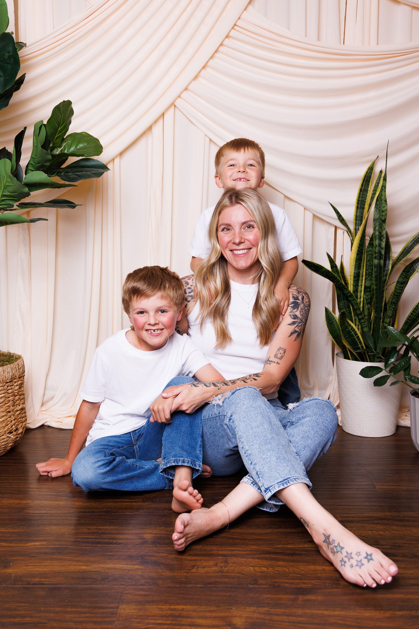 A smiling mother with tattoos sits on a hardwood floor with her two blonde sons in matching white tees and jeans.