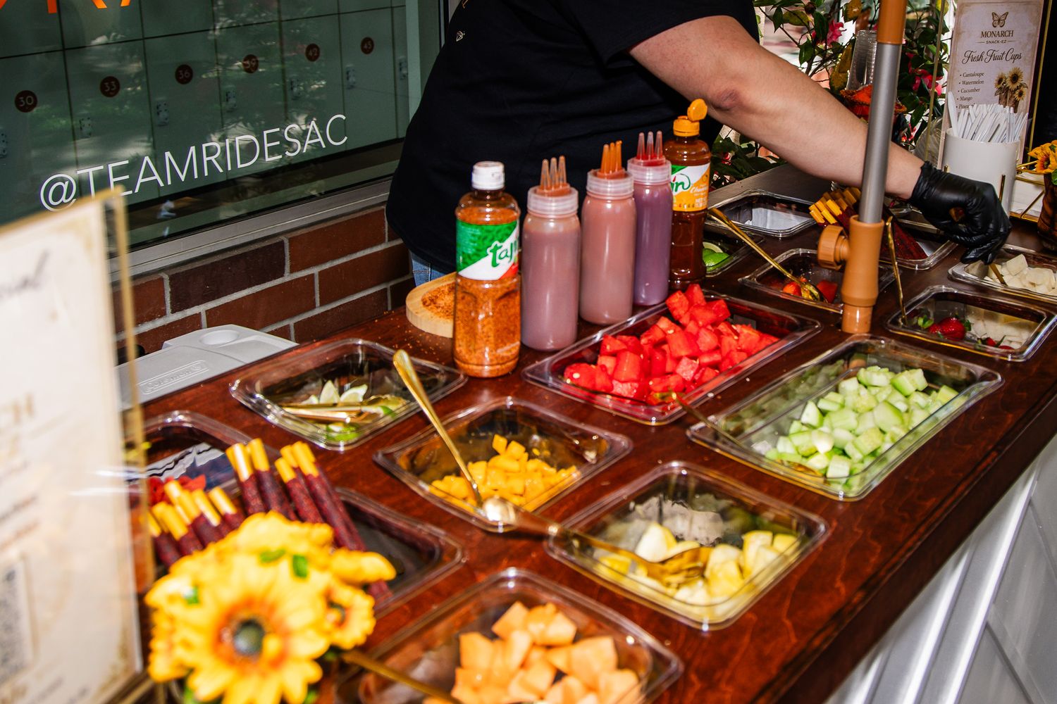 Fresh fruit and condiment bar setup with colorful toppings in wooden compartments and bottled sauces on display.