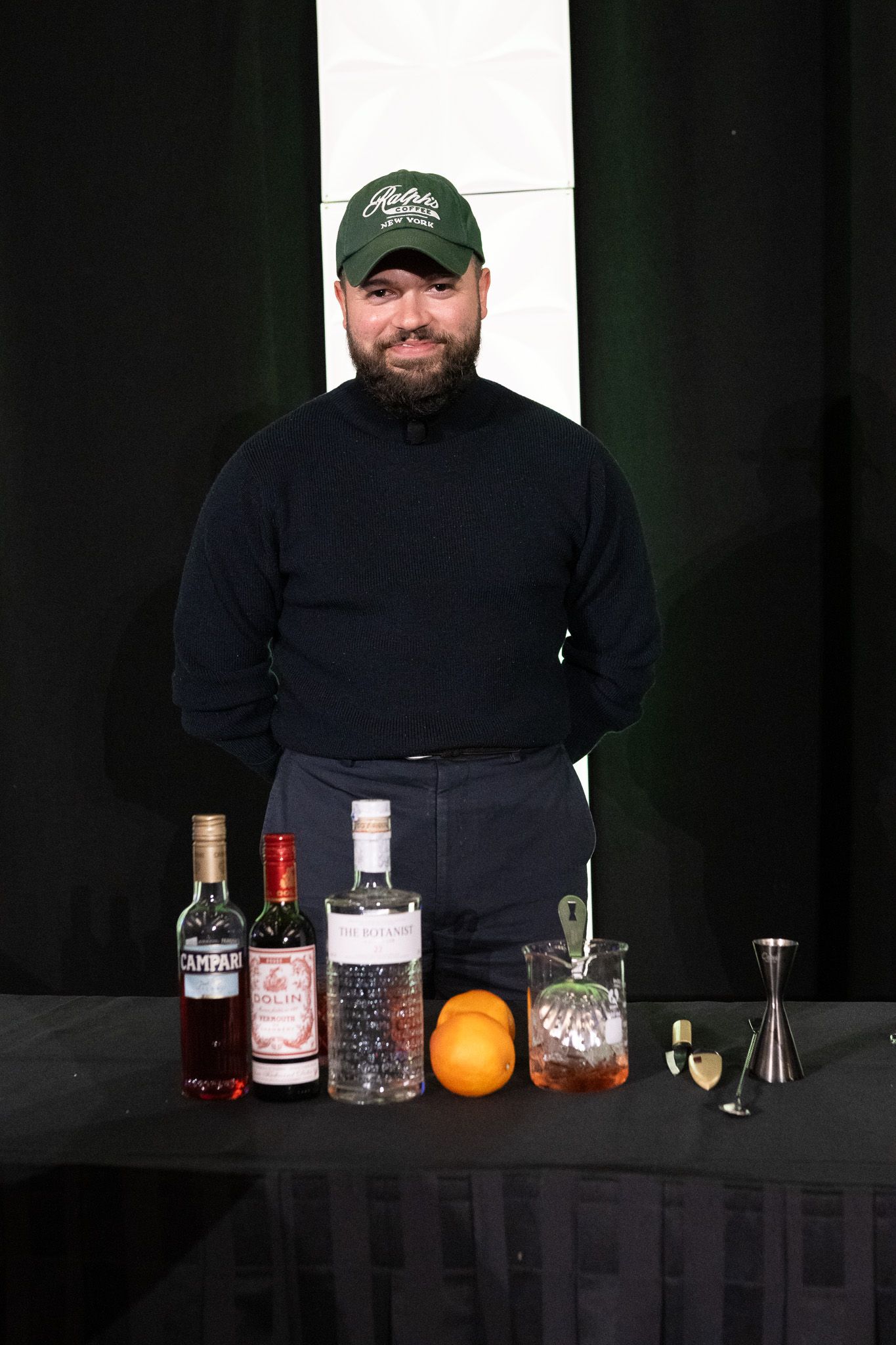 A bartender in dark clothing stands behind a table display of spirits, mixers and bar tools with an orange garnish.