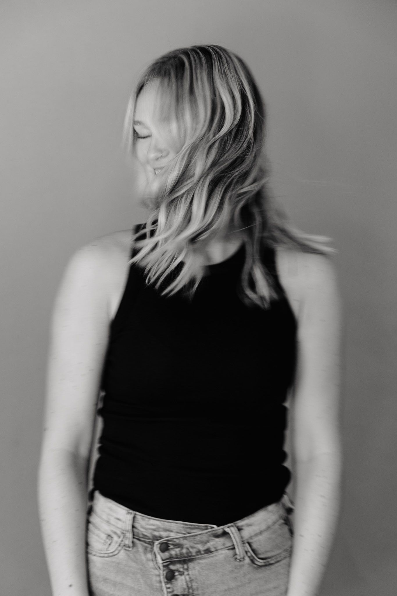 Black and white portrait of someone with light wavy hair wearing a tank top and jeans against a plain backdrop.