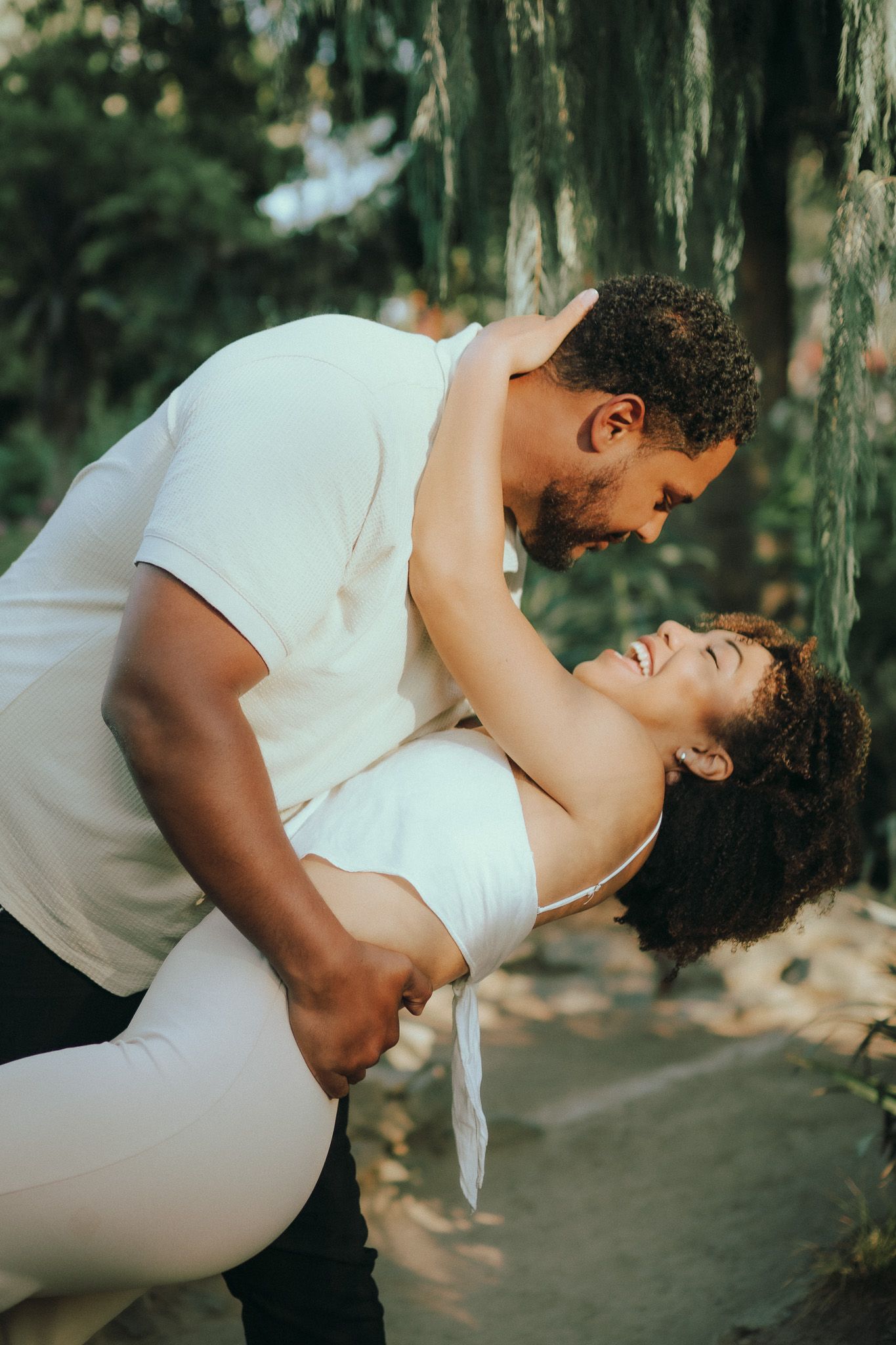 A romantic couple in white outfits share an intimate dance pose in a wooded outdoor setting.