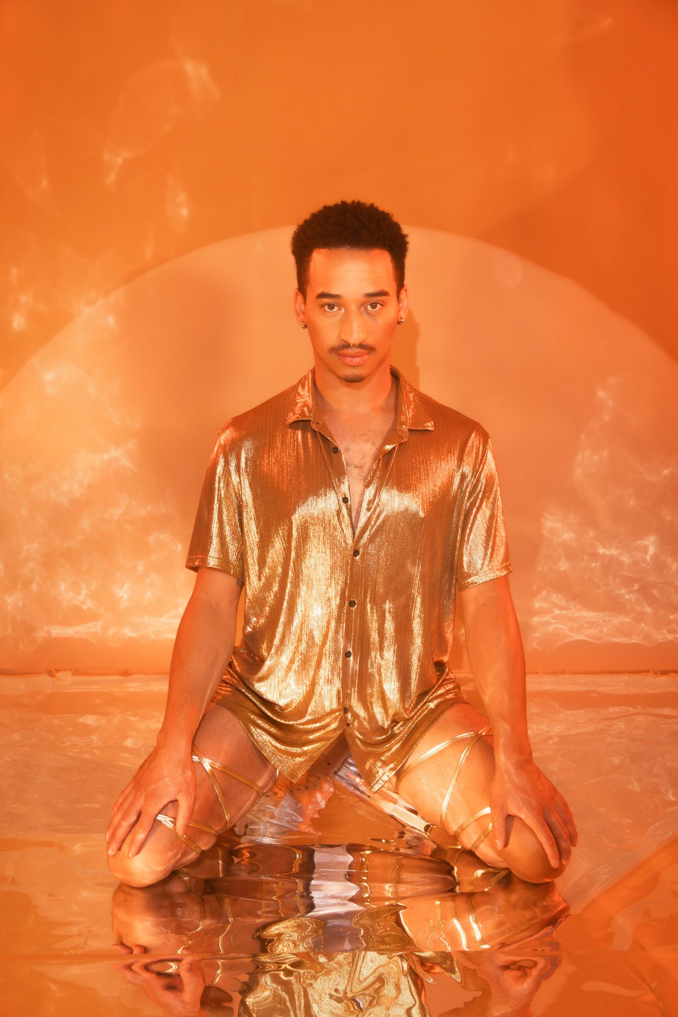 Model in metallic gold shirt poses in meditation position on reflective surface with orange lighting.
