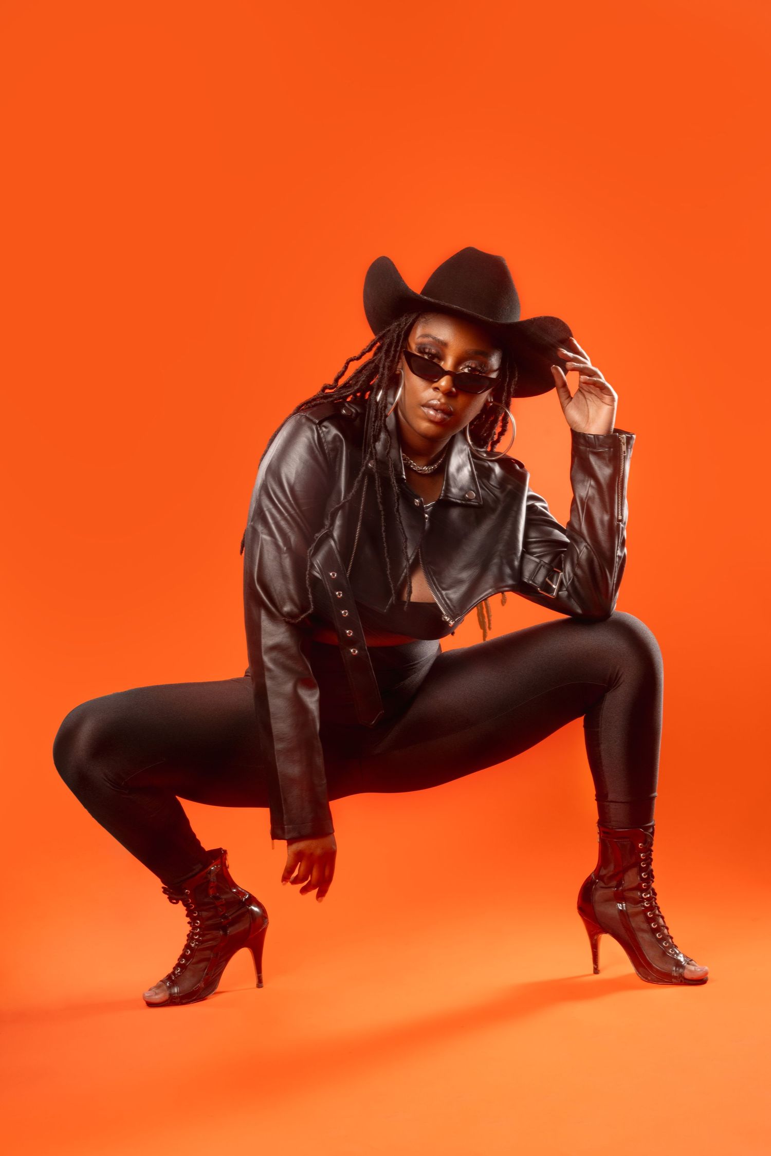 Fashion model in black leather jacket and boots strikes dramatic poses against vibrant orange background in photo series.