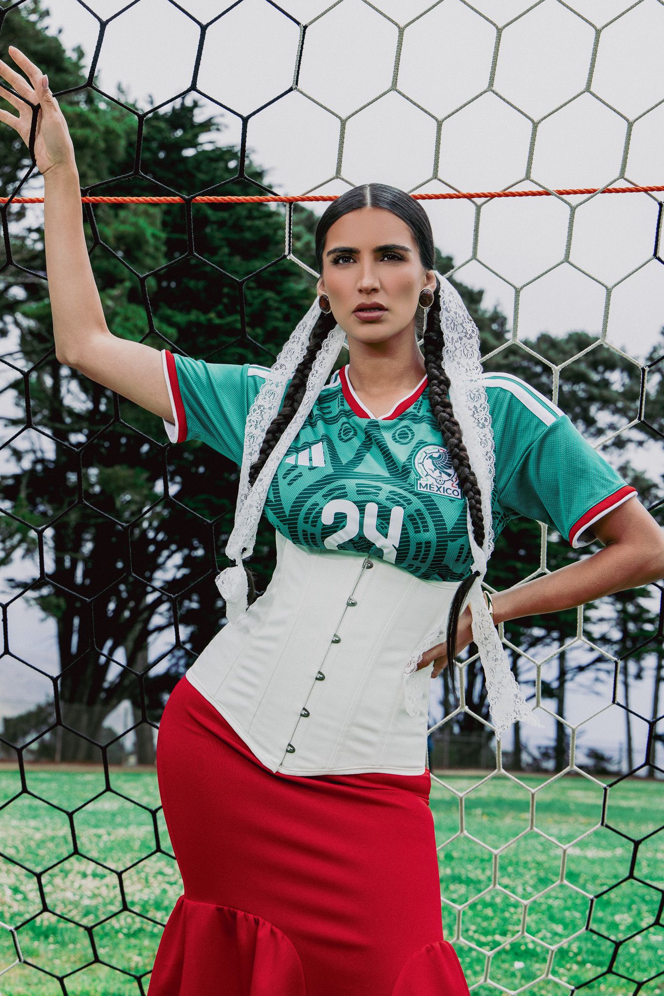 Woman in Mexico soccer jersey, white corset, and red skirt poses against a soccer goal net outdoors.