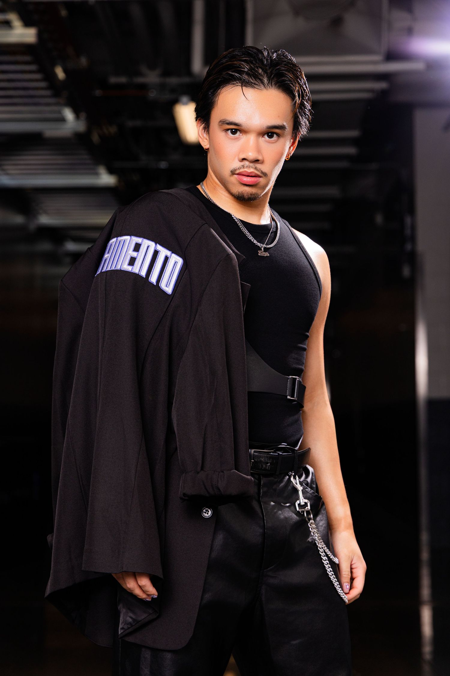 Young man in black streetwear outfit with oversized jacket, harness, and chain accessories poses confidently indoors.