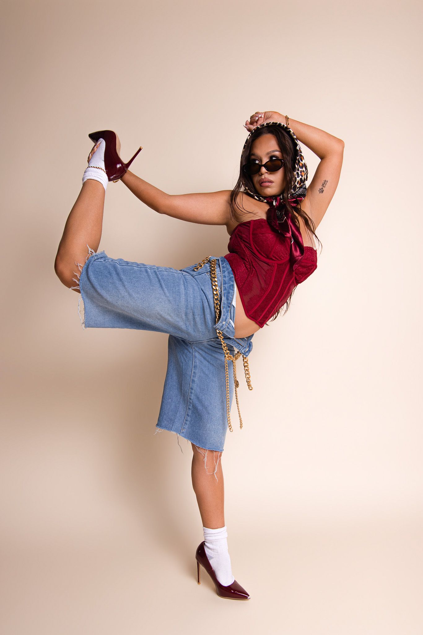 Woman in red top, denim skirt, and burgundy heels striking a high kick pose against a beige background.