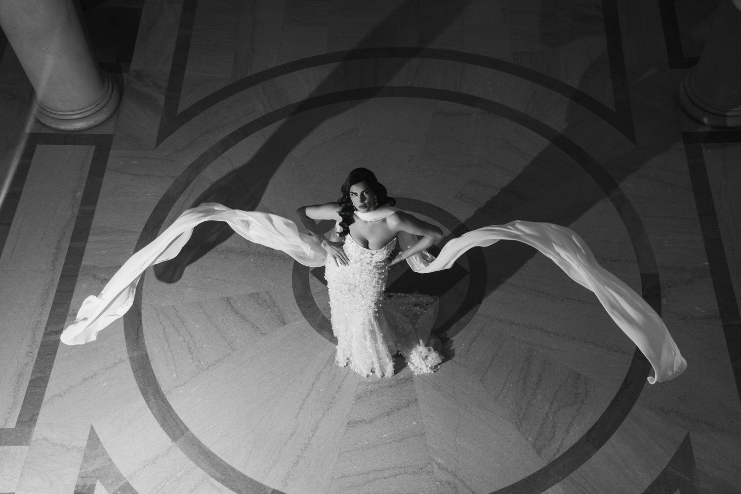 Aerial black and white photo of a bride in a gown with flowing fabric wings on ornate floor.