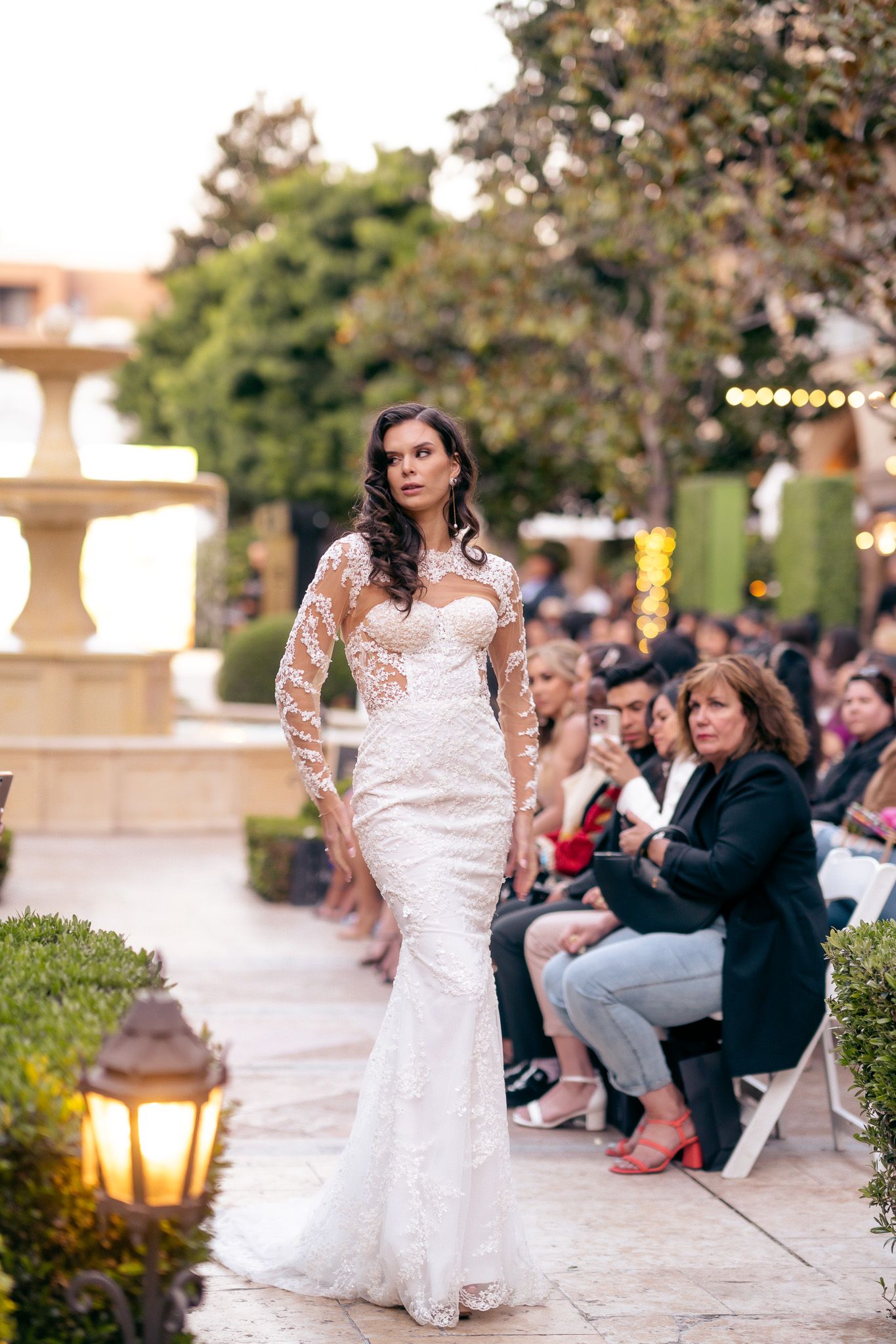 A bride in a lace wedding dress walks gracefully along an outdoor venue path at sunset with guests seated nearby.