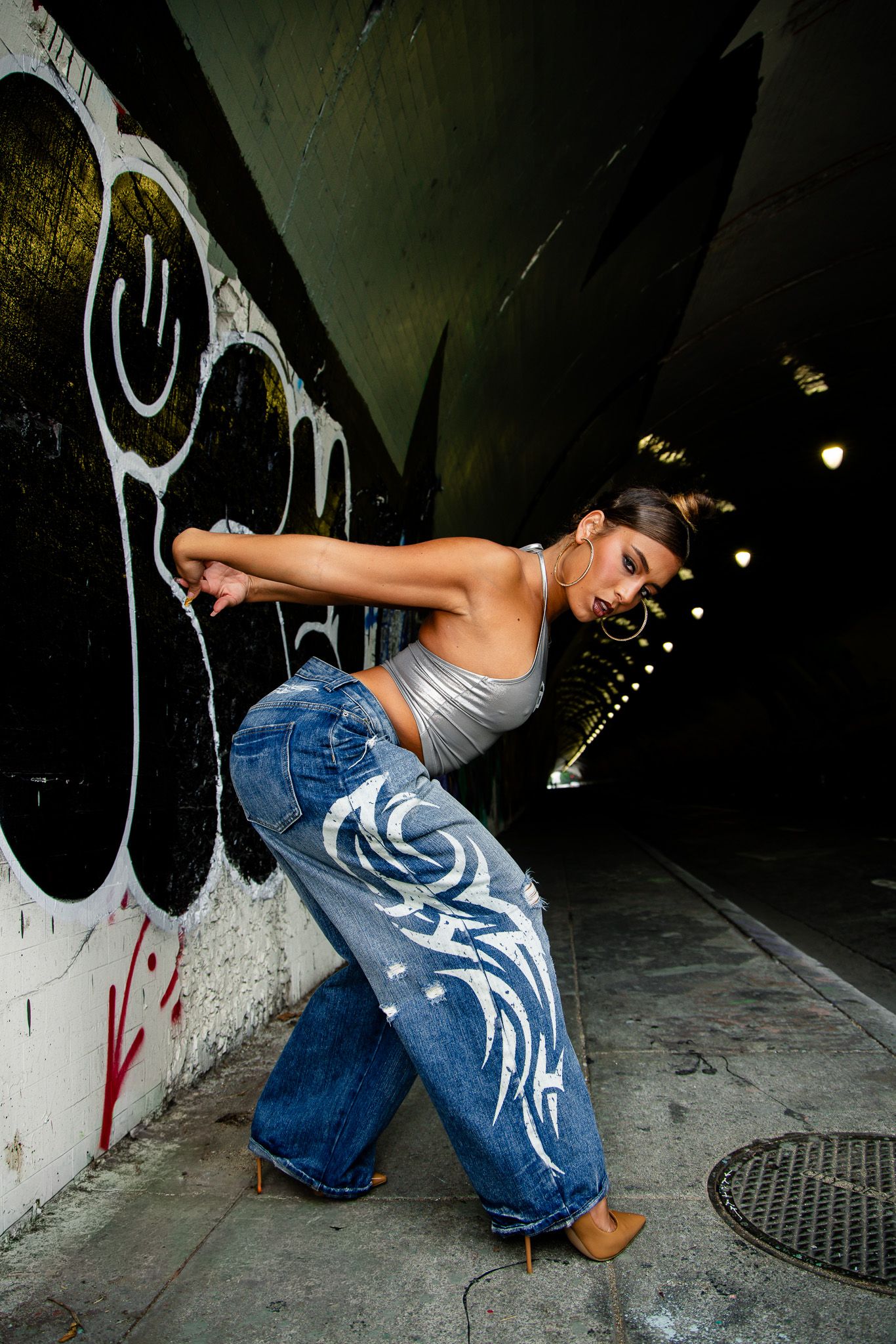 Urban dancer in white top and baggy jeans performs dynamic moves against graffiti wall in dark tunnel.