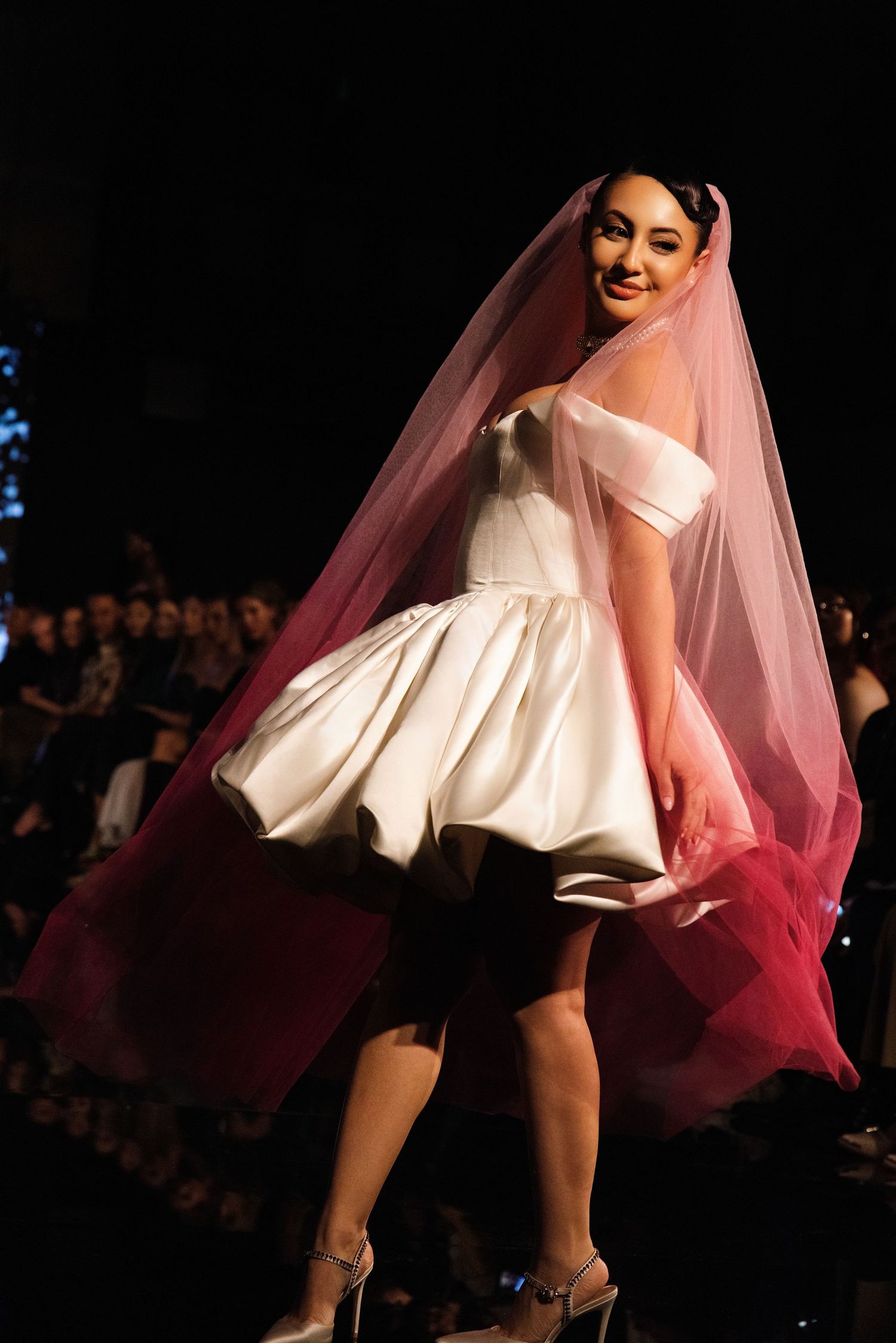 A model walks the runway in a short white wedding dress with off-shoulder design and flowing pink veil at a fashion show.