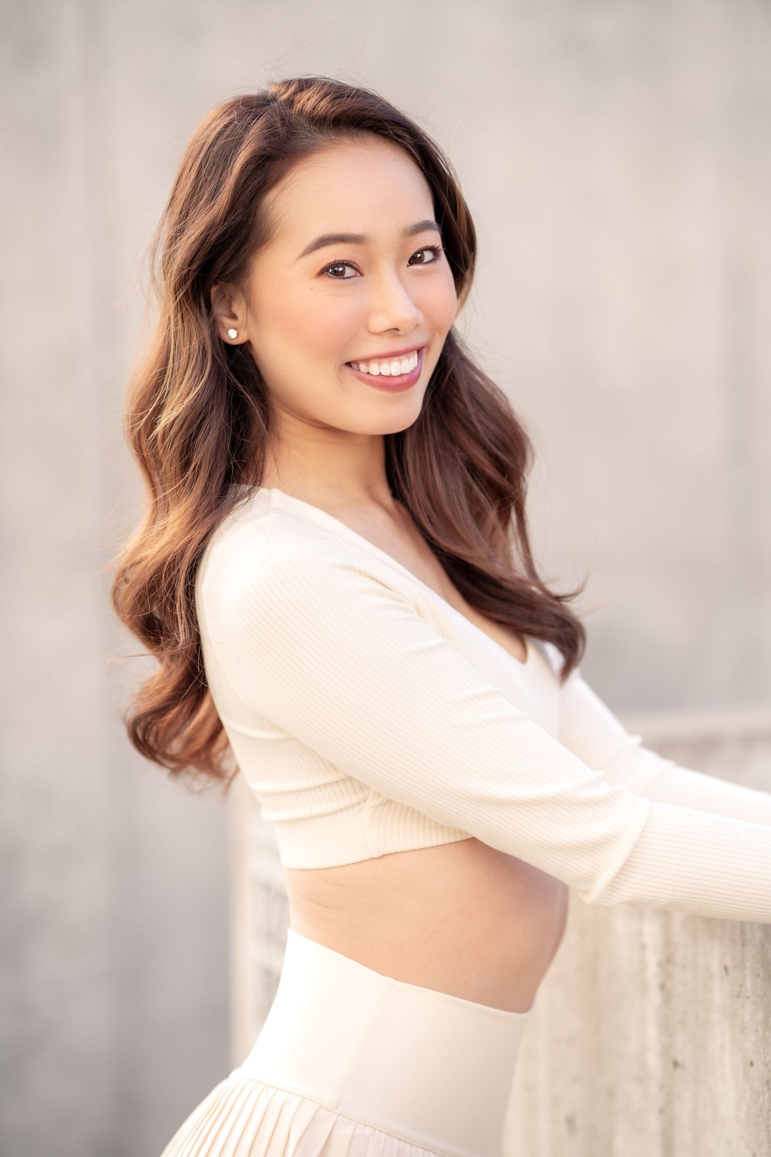 Woman with long wavy brown hair poses in white crop top and skirt against gray wall.