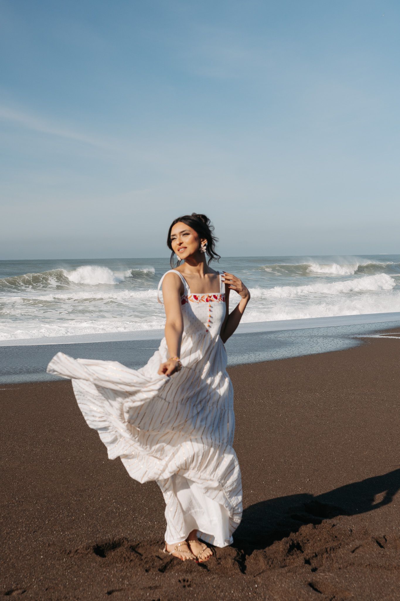 A person in a flowing white dress stands against the dramatic backdrop of a beach with rolling waves and dark sand.