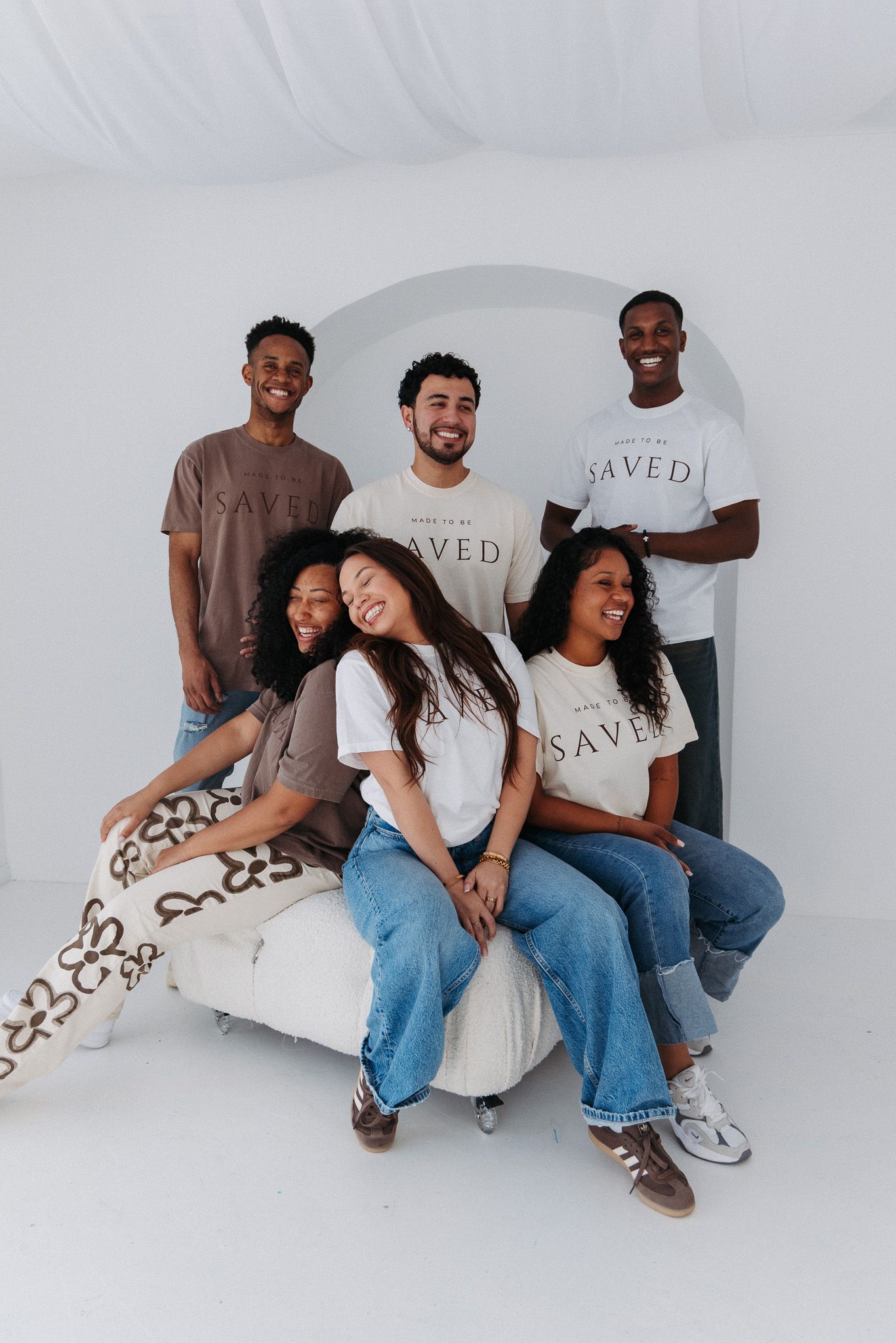 Group relaxing on white furniture wearing coordinated outfits of jeans and SAVED branded shirts.