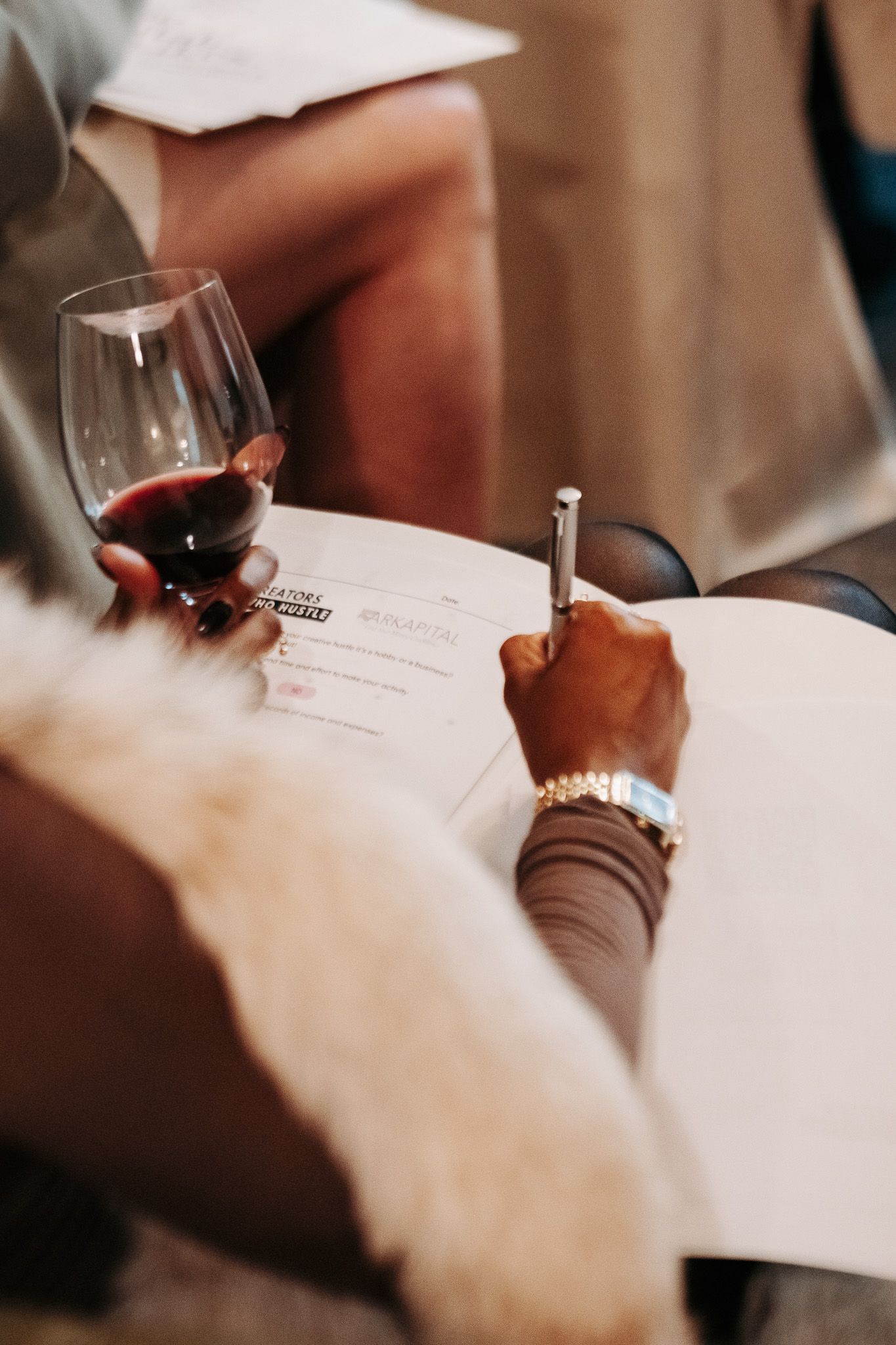 Close-up of hands writing notes on white paper with wine glasses visible.