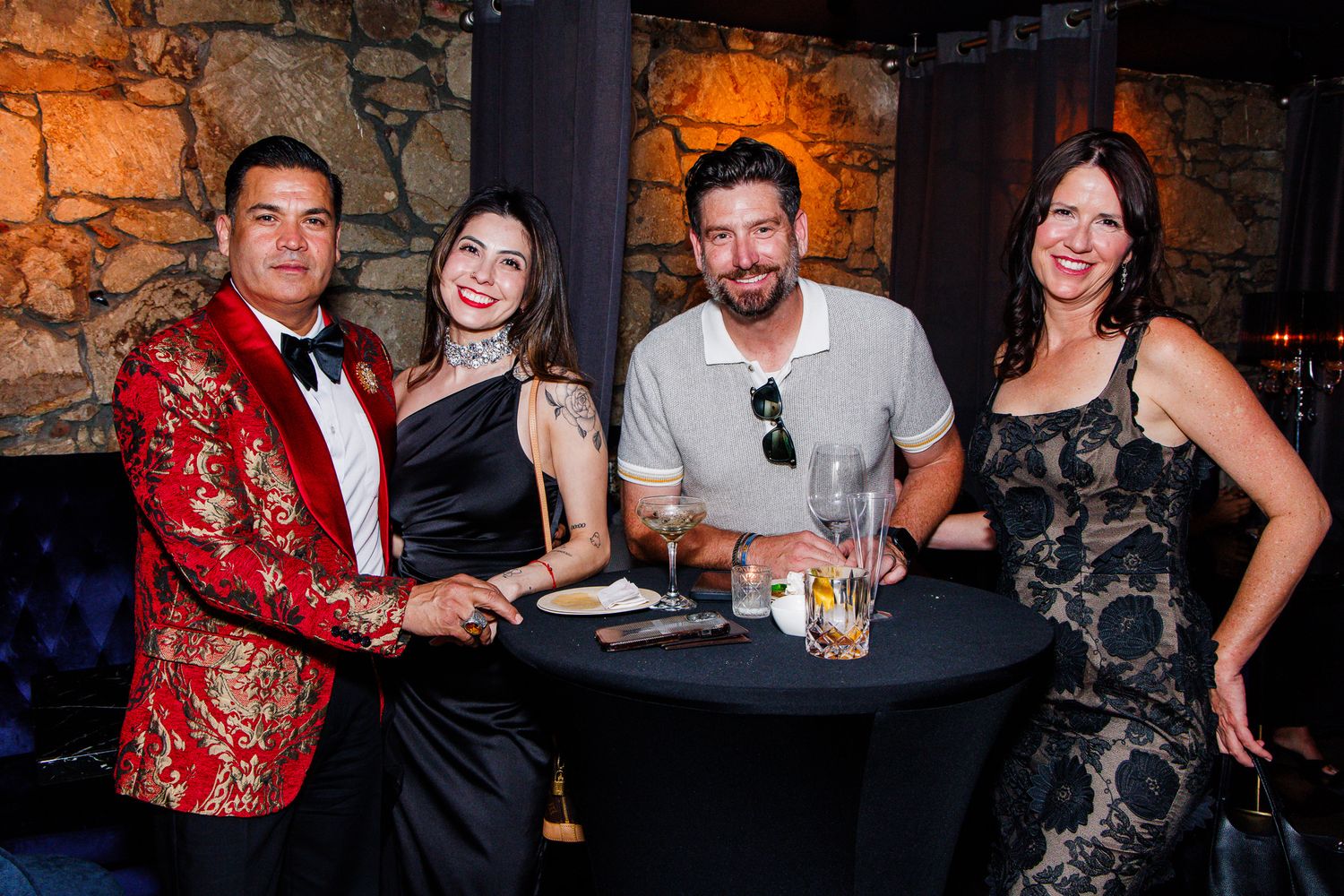 Four elegantly dressed guests smile together at a cocktail table during an upscale evening event.