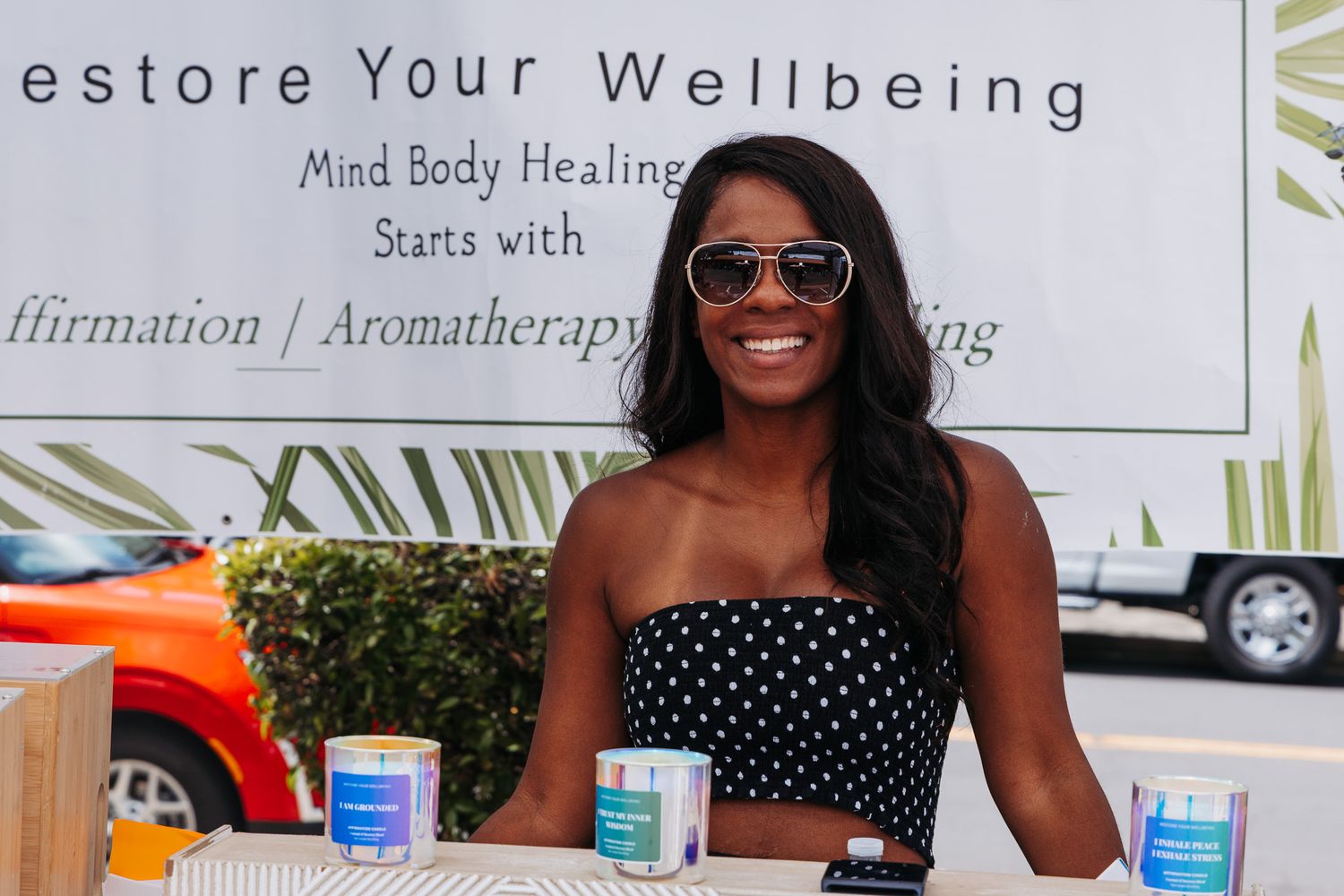 A smiling woman in a polka dot dress stands at a wellness booth promoting mind body healing products.