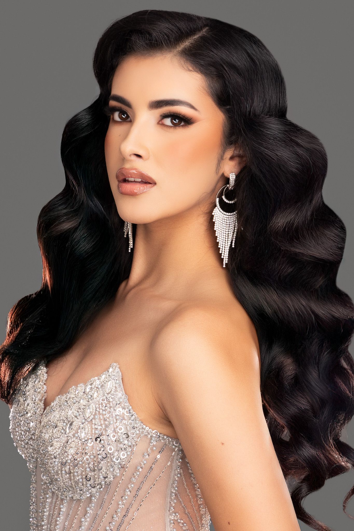 A model with dark wavy hair and glamorous makeup poses in a crystal-embellished strapless gown against a gray background.