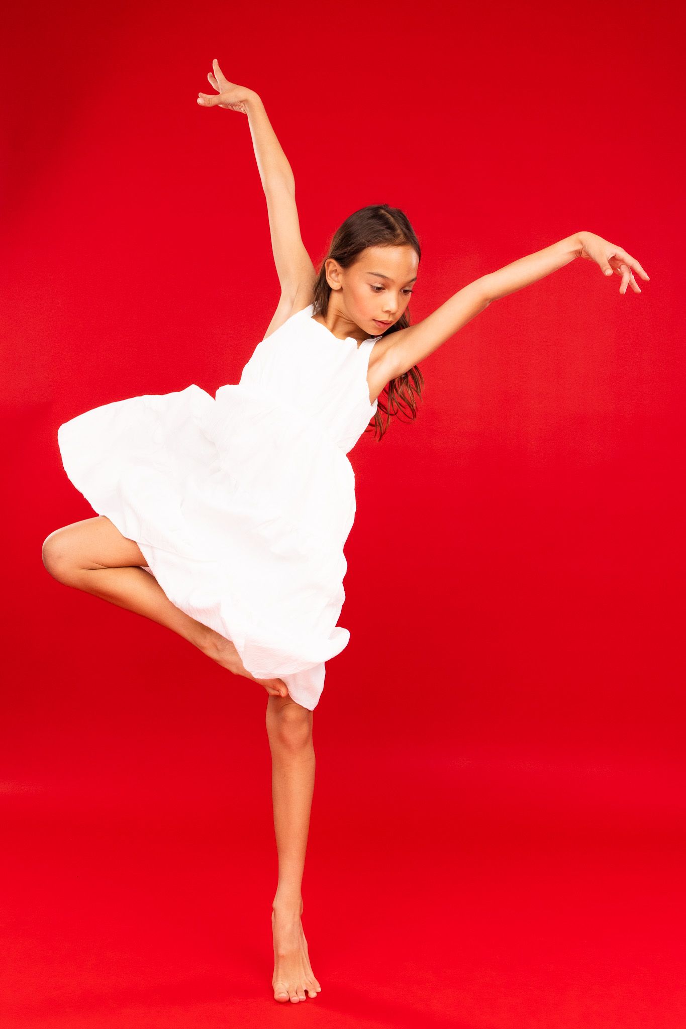 Young dancer in flowing white dress performs graceful movements against vibrant red backdrop.
