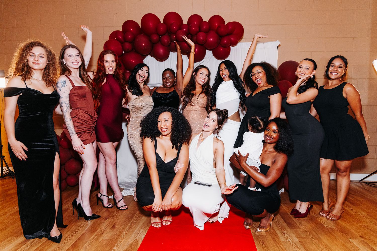 Group of women in elegant dresses posing together in front of red balloon arch at a celebration event.