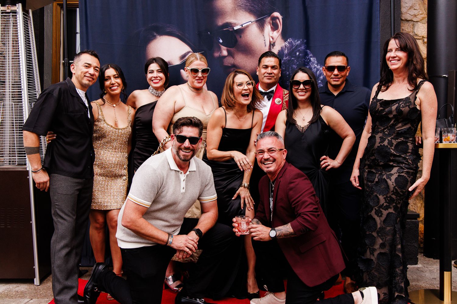Group of elegantly dressed people posing together at a red carpet event in front of a movie poster backdrop.