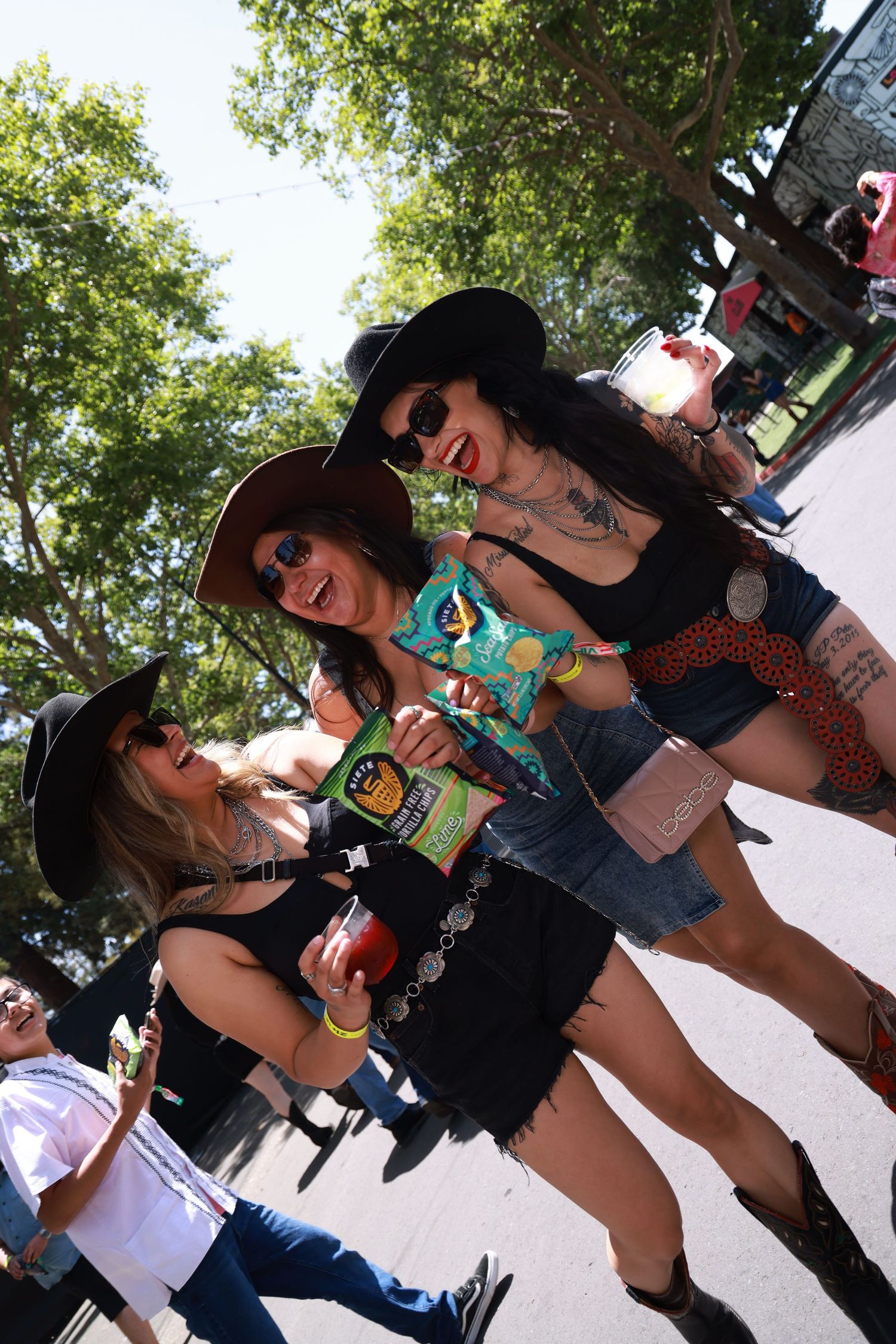 Three women in black cowboy hats smile and pose outdoors at a festival on a sunny day.