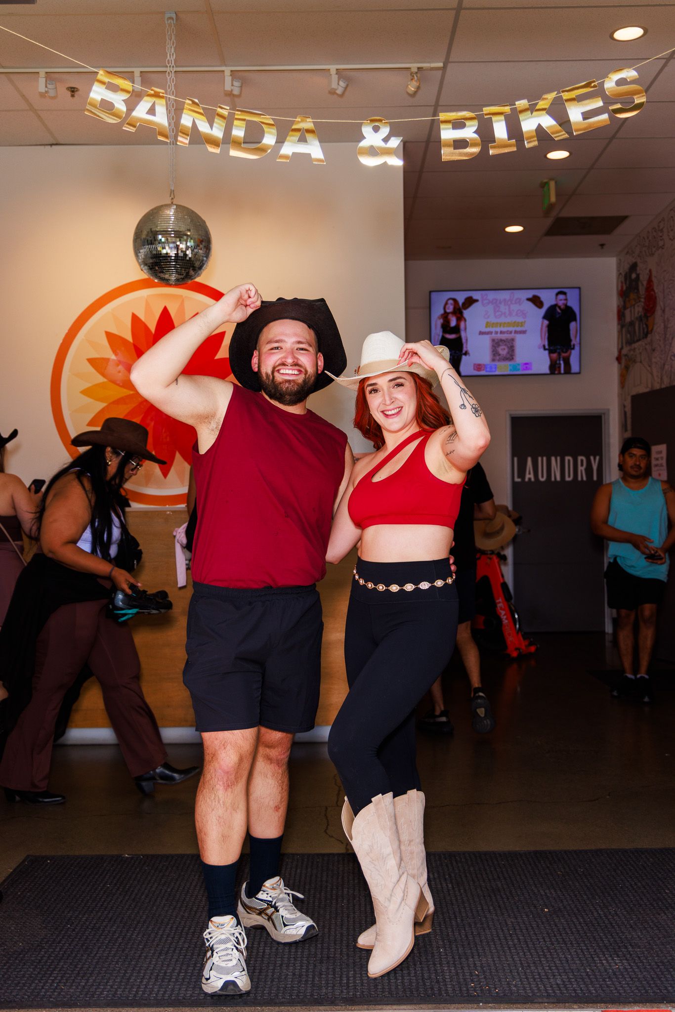 Two people in cowboy hats pose at a Banda & Bikes event inside a fitness studio with a disco ball.
