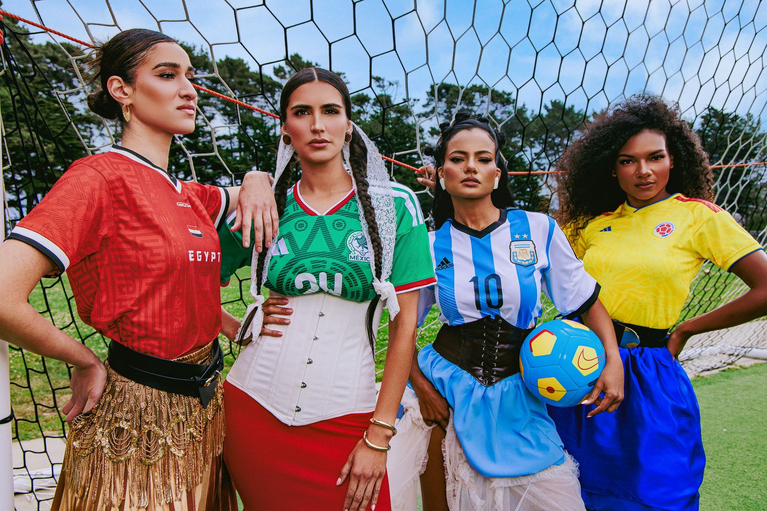 Four women in colorful soccer jerseys pose together on a field near a goal net, one holding a soccer ball.