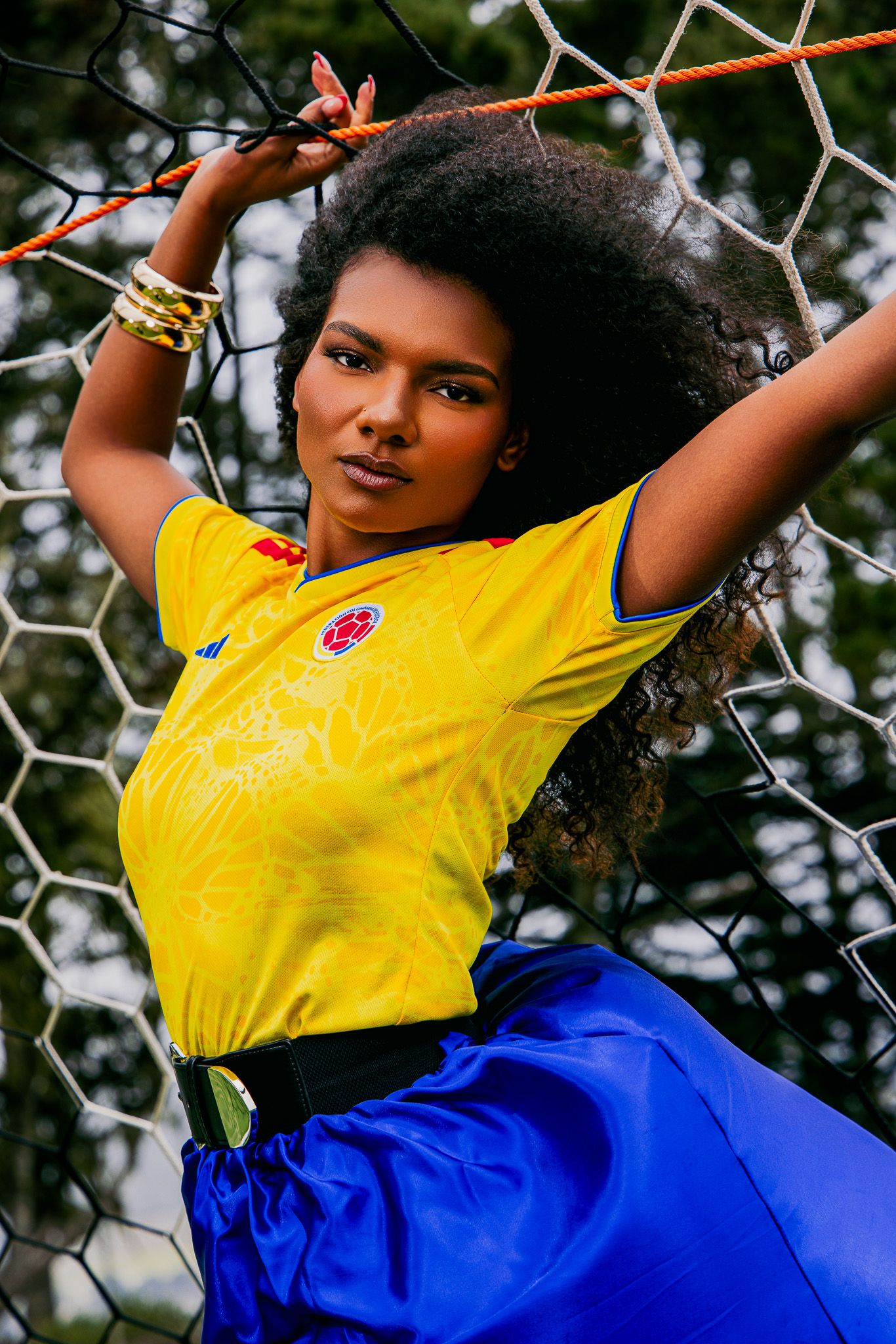 Young woman in Colombia soccer jersey and blue skirt posing against a goal net outdoors.