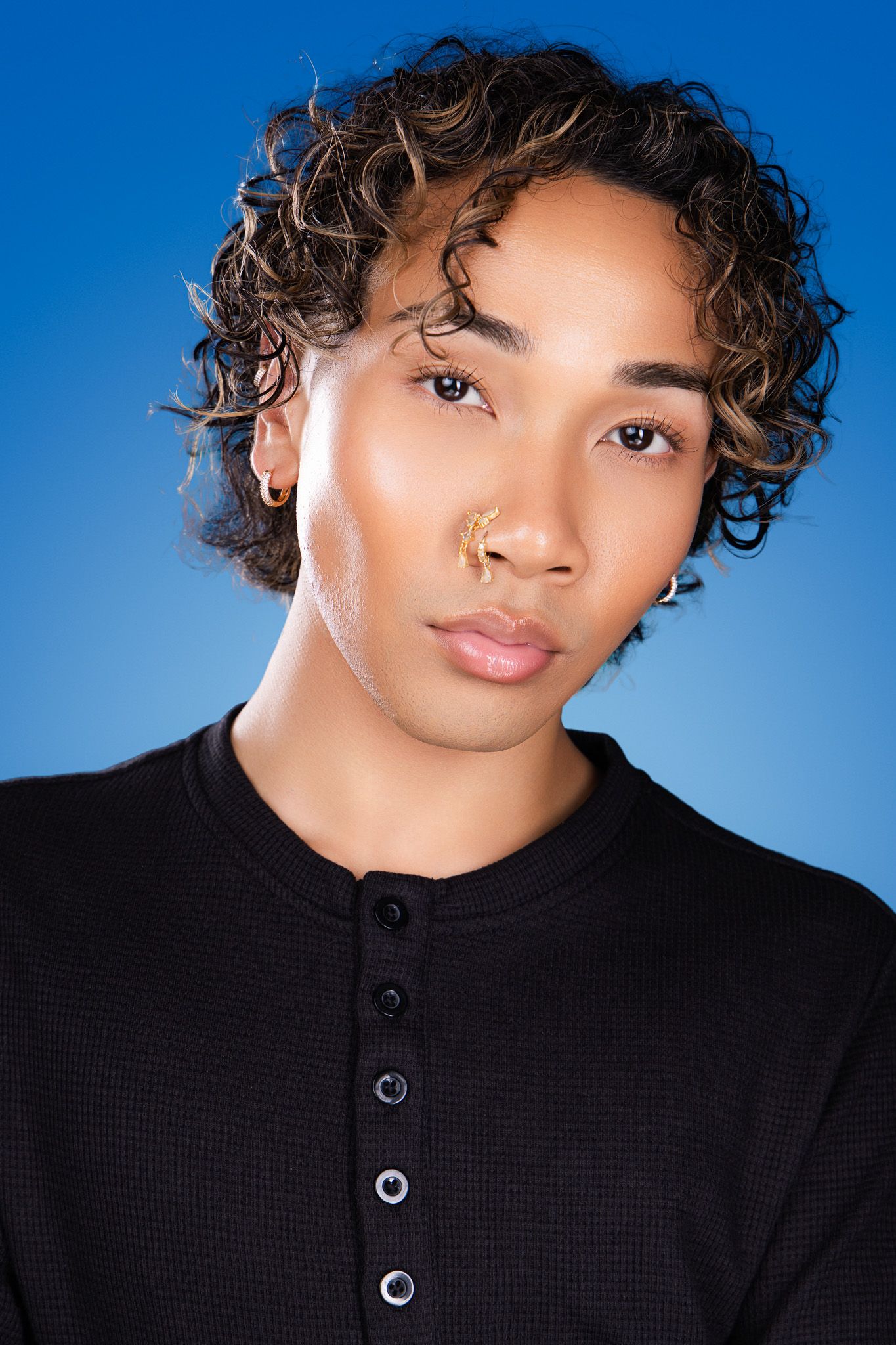 Person with curly highlighted hair and gold nose ring in black henley shirt posing against blue backdrop.