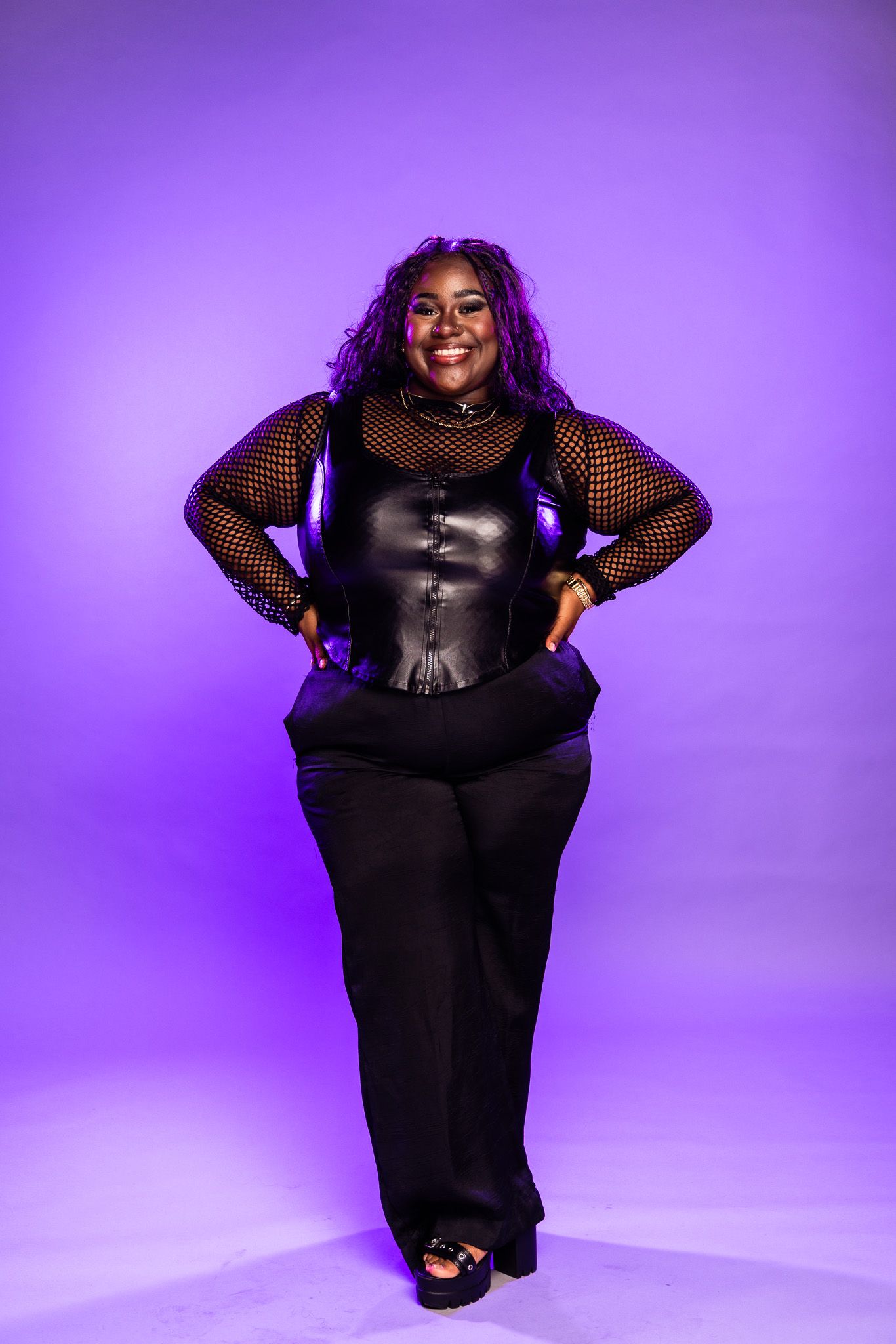 Model in black mesh and leather ensemble poses confidently against vibrant purple studio background.