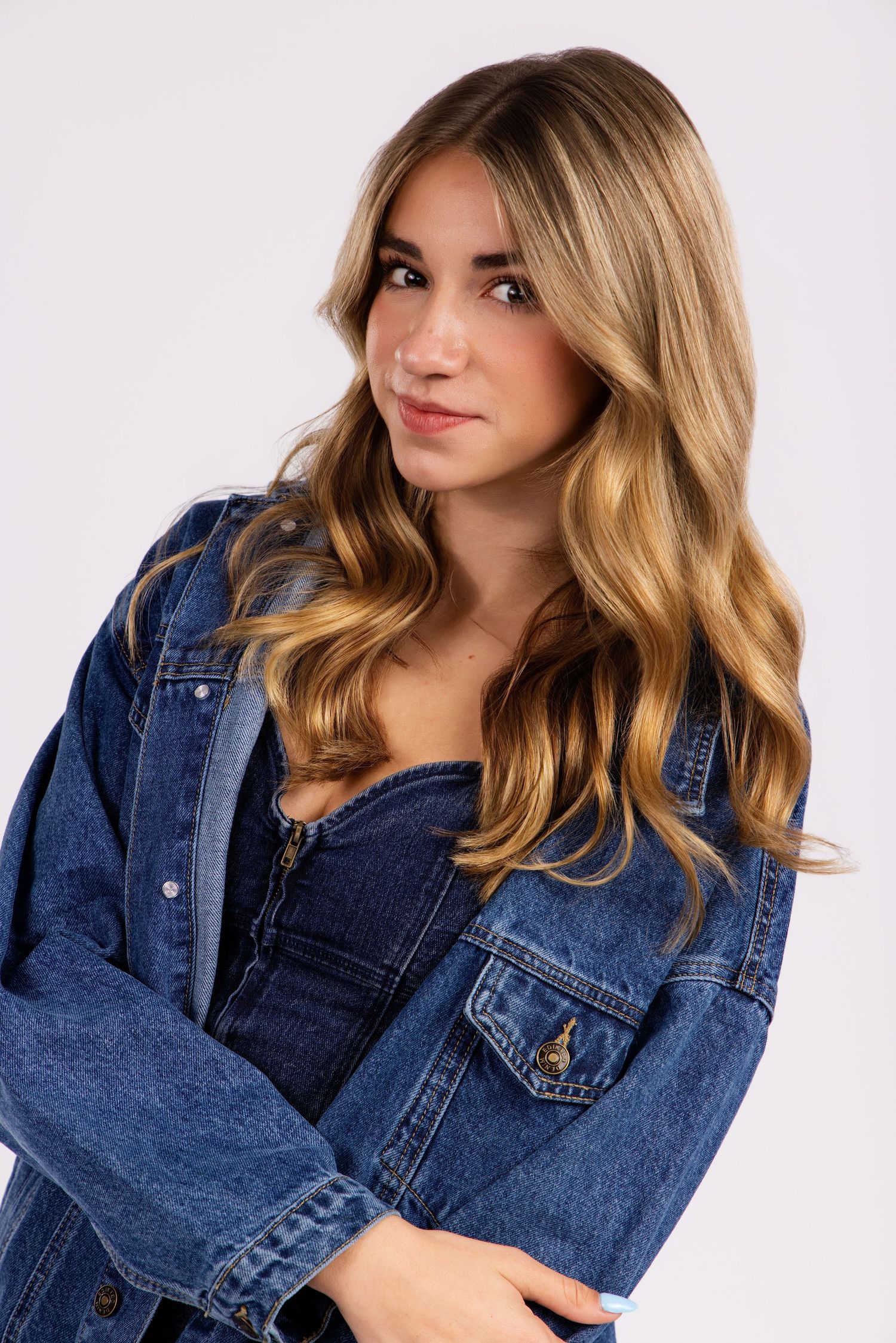 Model wearing denim jacket and top with wavy blonde hair against light background in studio portrait.
